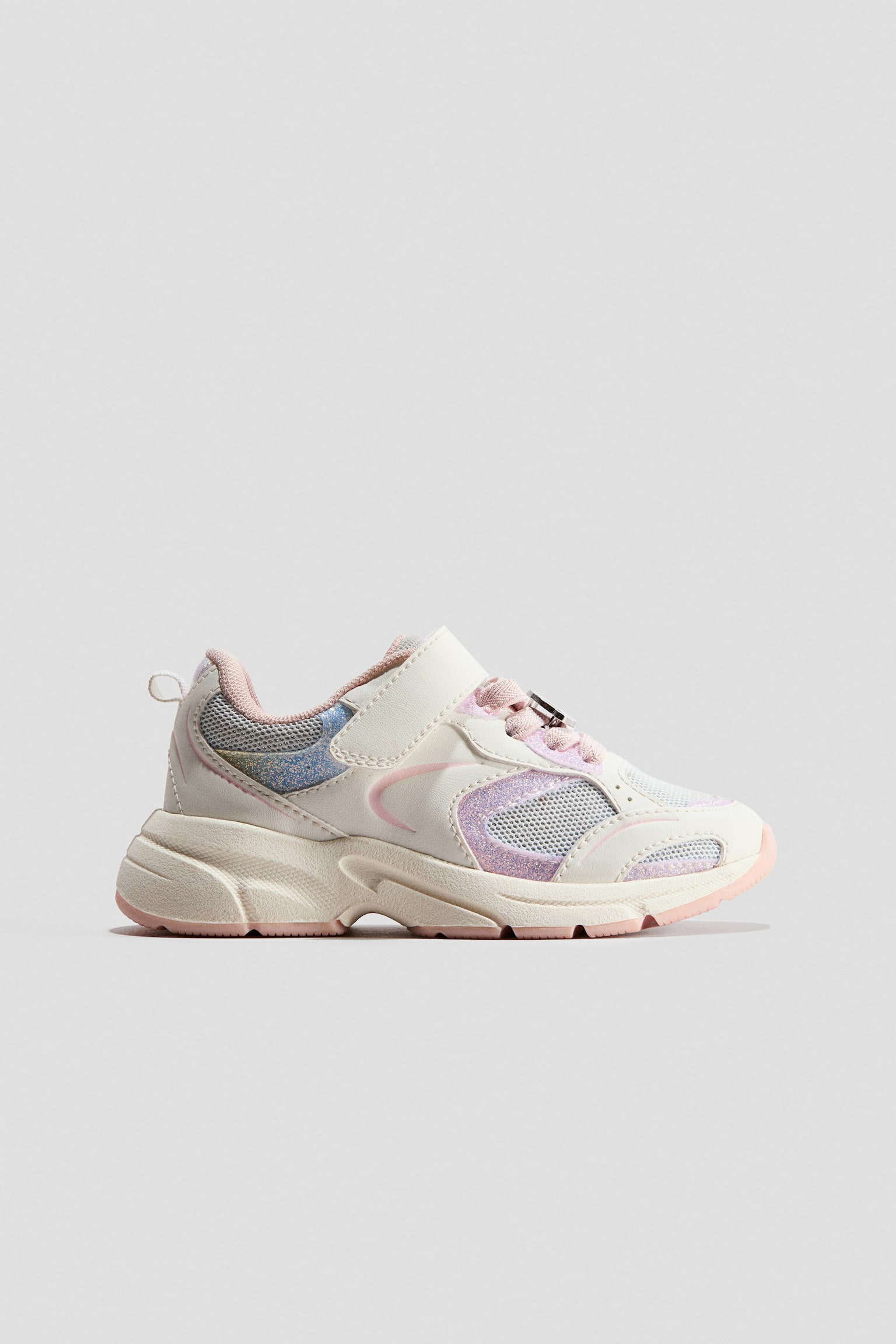 Sneakers are shown in a side profile view, featuring an off-white upper with glittery pink and iridescent blue panels alongside breathable mesh sections. The design incorporates a hook-and-loop strap closure over pink laces, a heel pull tab, and a chunky textured midsole with a pink outsole.