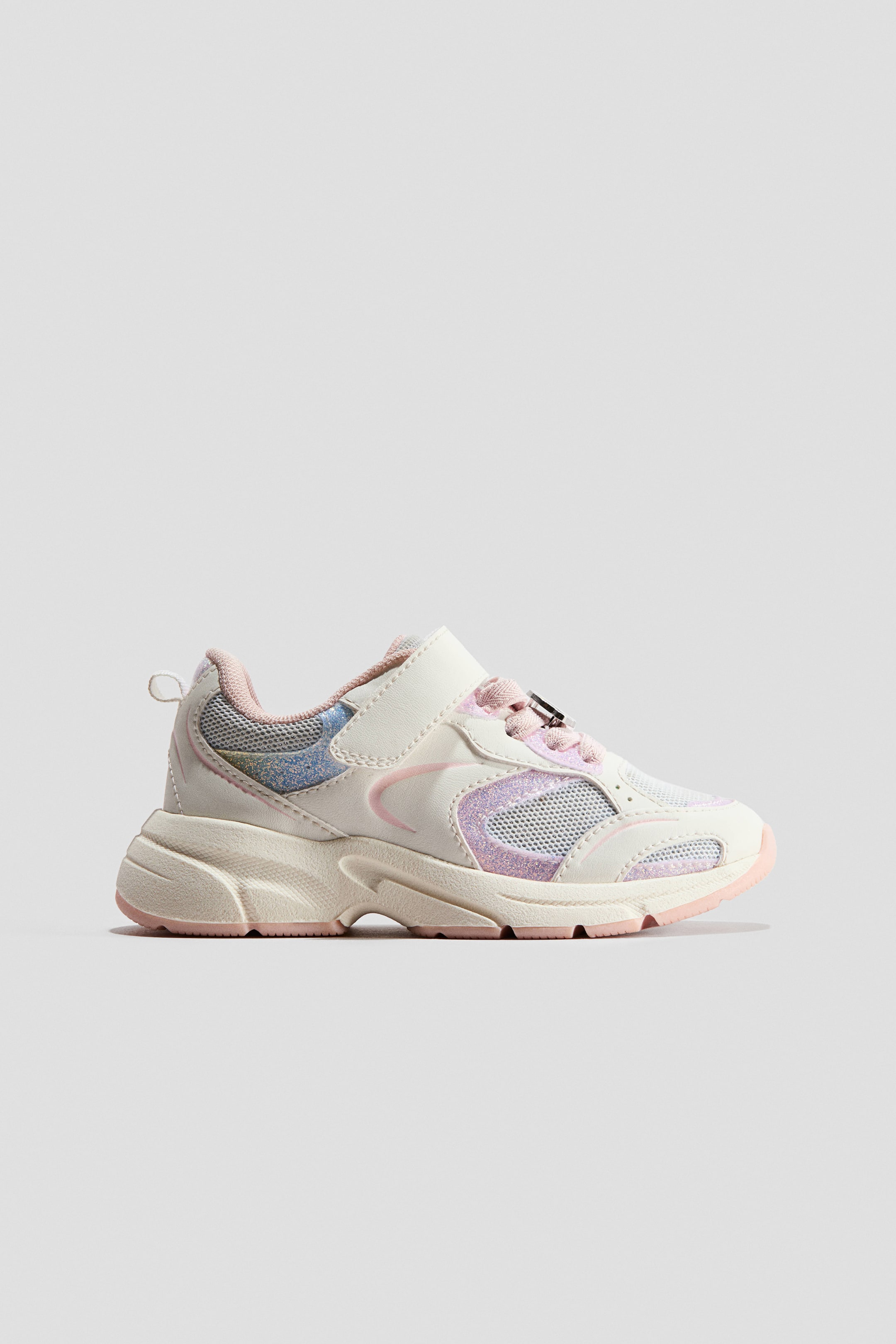 View larger image: Sneakers are shown in a side profile view, featuring an off-white upper with glittery pink and iridescent blue panels alongside breathable mesh sections. The design incorporates a hook-and-loop strap closure over pink laces, a heel pull tab, and a chunky textured midsole with a pink outsole.