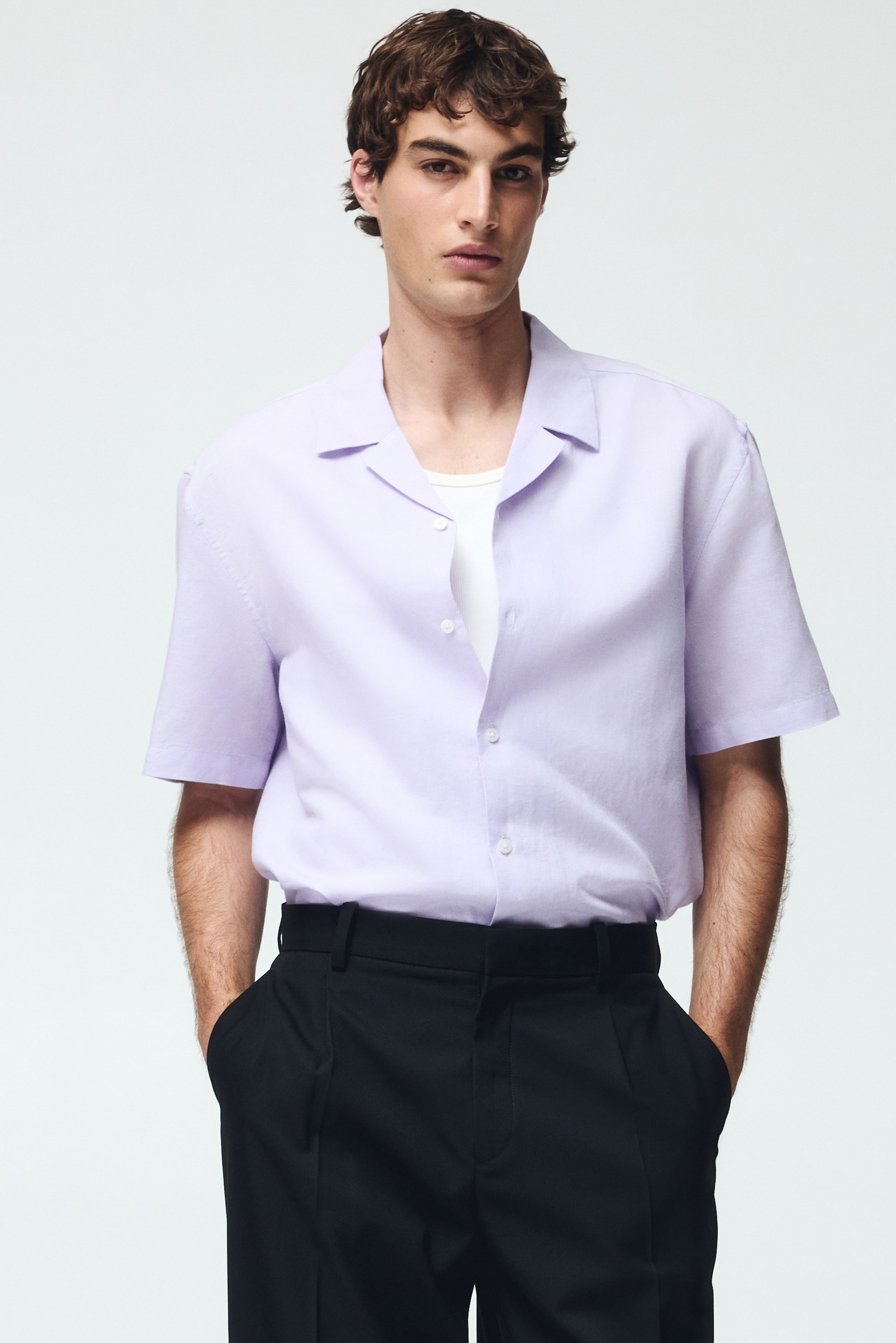 View larger image: A light purple short-sleeved shirt features a Cuban collar and white front buttons. It is styled open over a white ribbed undershirt, with its short sleeves ending just above the elbows, and is neatly tucked into black trousers.