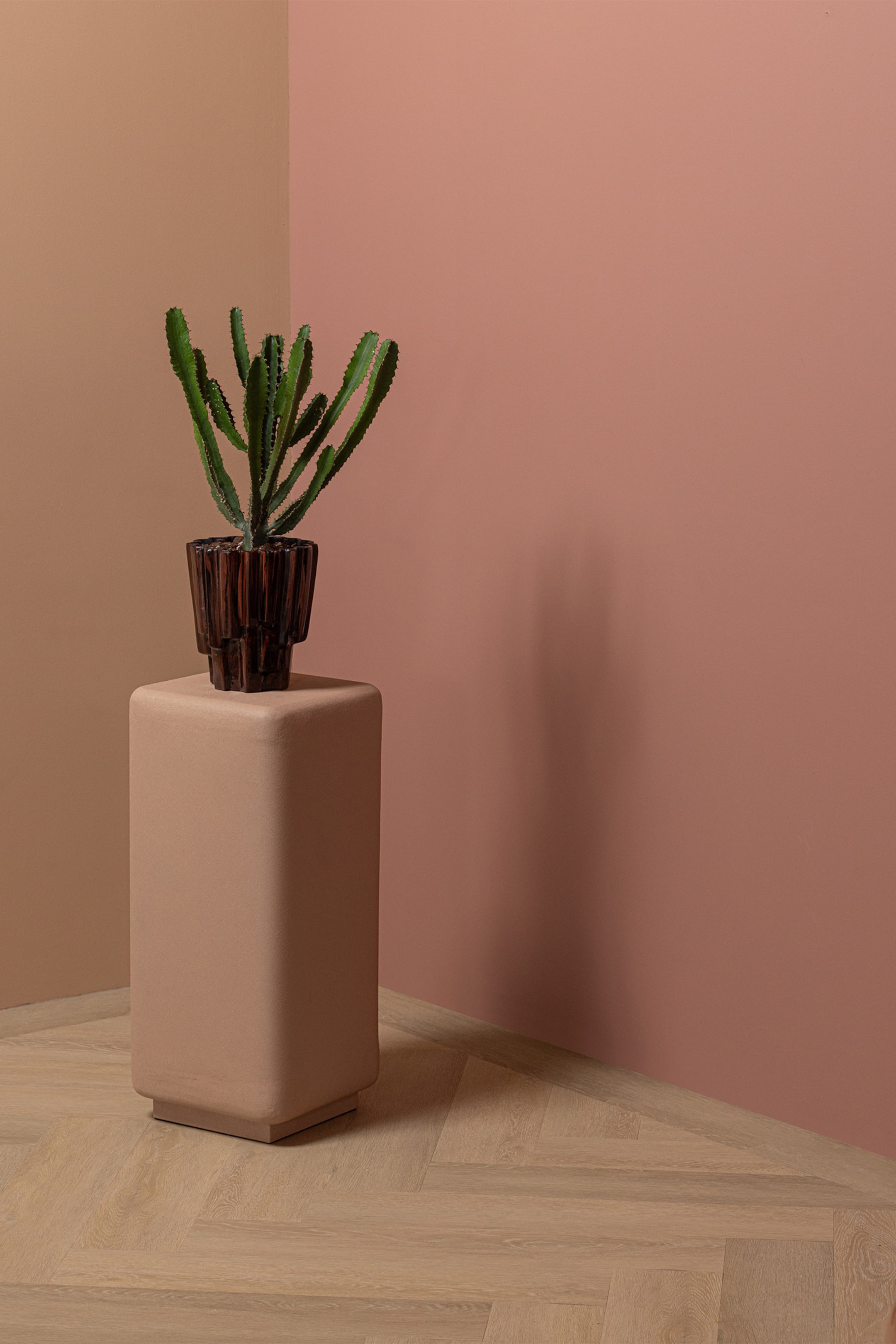 Agrandir l'image: Un cactus vert à plusieurs tiges pousse dans un pot marron foncé à rainures, doté d'une base en escalier. Ce pot repose sur un haut piédestal carré aux bords arrondis, fini dans une teinte marron clair.