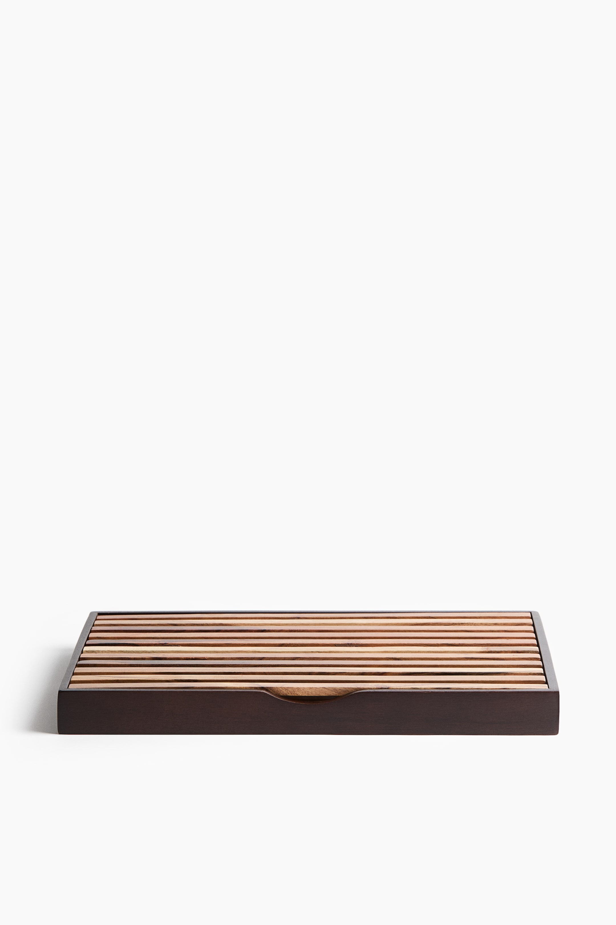 View larger image: A wooden breadboard featuring a removable grate on its top surface, composed of parallel slats in varying light and dark wood tones. The board also presents a dark wooden crumb-catching tray underneath, visible from the front with a curved access cutout.