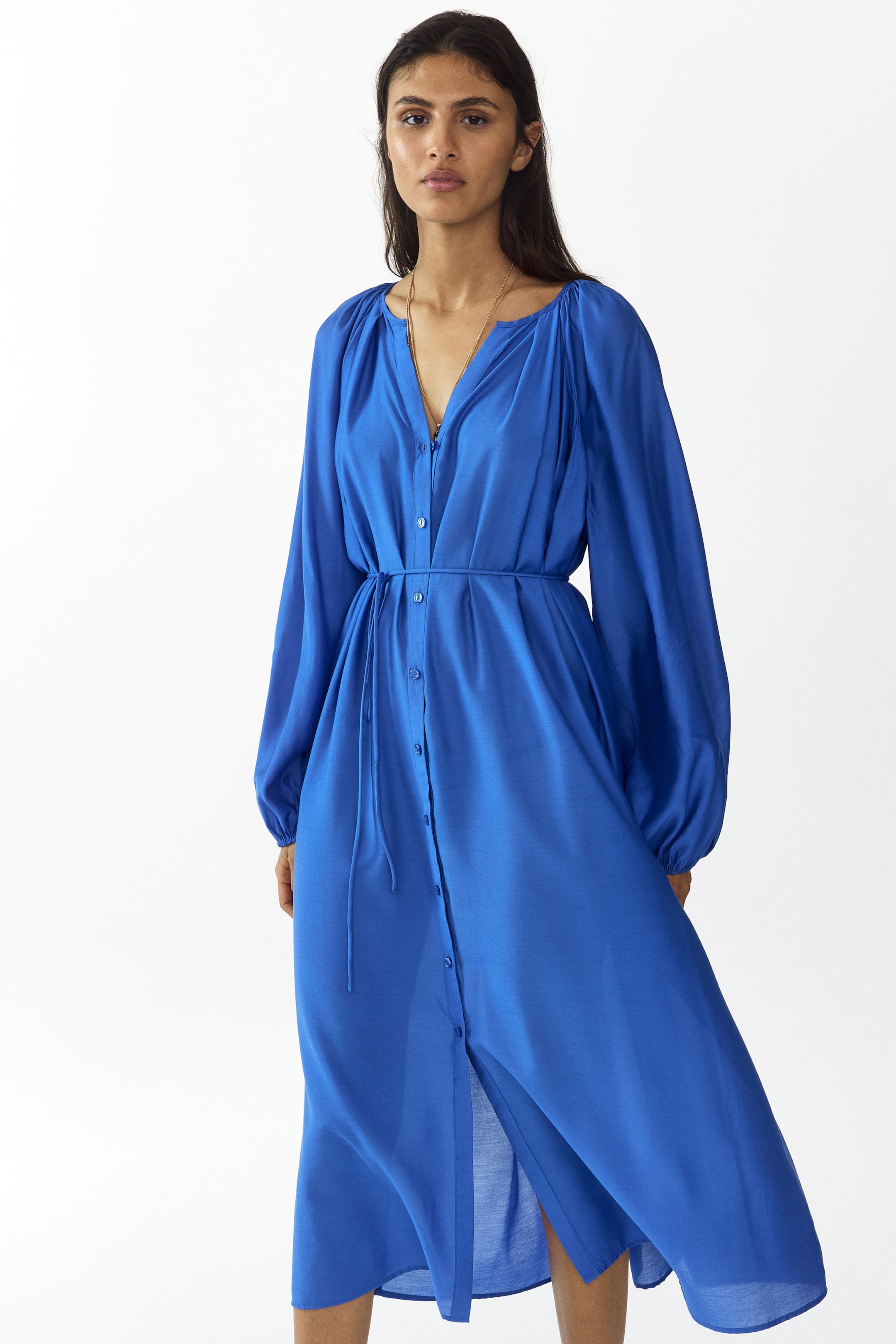 Größeres Bild ansehen: Das blaue Midikleid zeichnet sich durch einen V-Ausschnitt, lange voluminöse Ärmel mit elastischen Bündchen und eine vollständige Knopfleiste vorne aus. Ein Bindegürtel aus dem gleichen Stoff betont die Taille, während die unteren Knöpfe offen bleiben und so einen vorderen Schlitz schaffen.