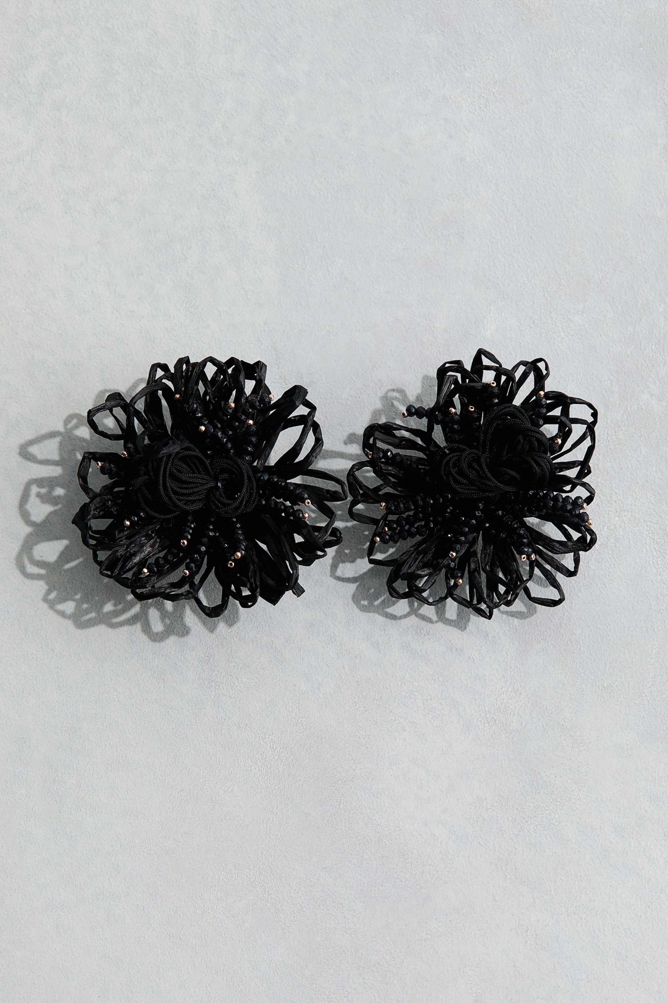 View larger image: Two black hair accessories are lying side by side, each featuring a central coiled elastic surrounded by ruffled, crinkled fabric strips and small embedded gold beads.