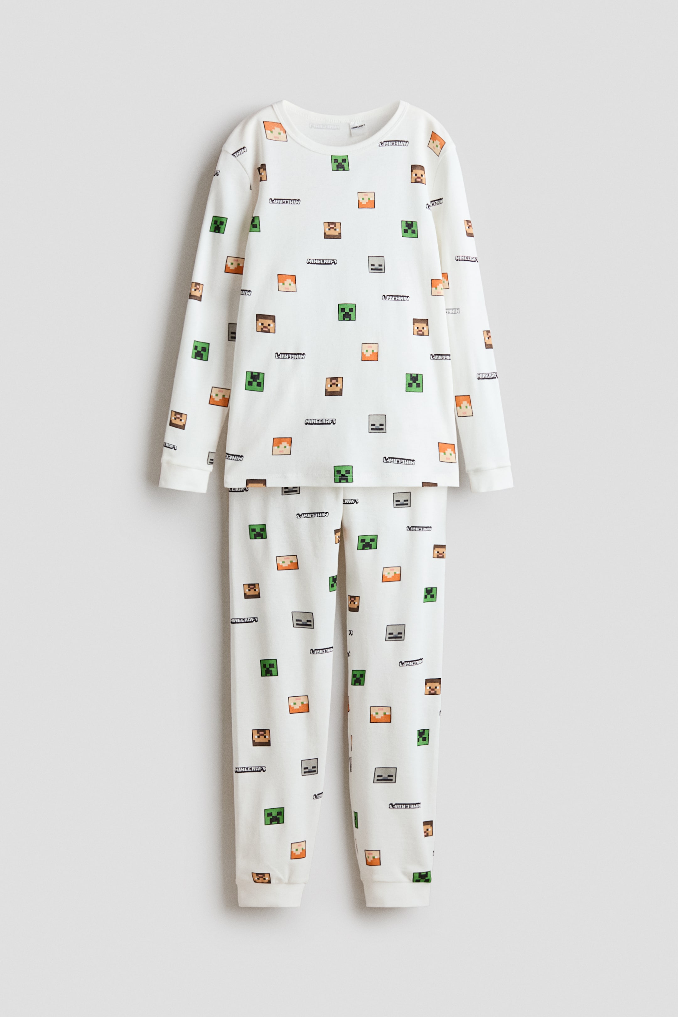 View larger image: A white two-piece pajama set is laid flat, displaying a long-sleeved top and matching long pants. The set features a repeating pattern of pixelated Minecraft character faces and the "MINECRAFT" logo. The top is crafted with a crew neck, and the pants are styled with elasticated cuffs, complementing the top's cuffs.