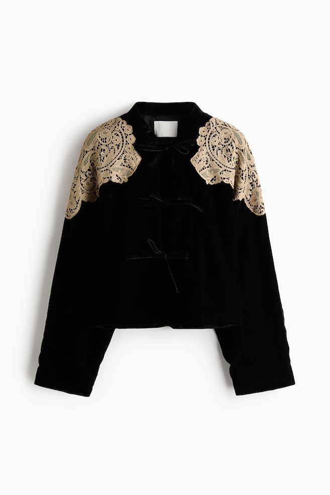 Una chaqueta de terciopelo negro vista de frente, que muestra delicadas aplicaciones de encaje beige dispuestas sobre los hombros y la parte superior de los brazos. La chaqueta presenta un cuello mao bajo, mangas largas rectas y tres cierres de lazo de cinta de terciopelo en la parte delantera central. - 1