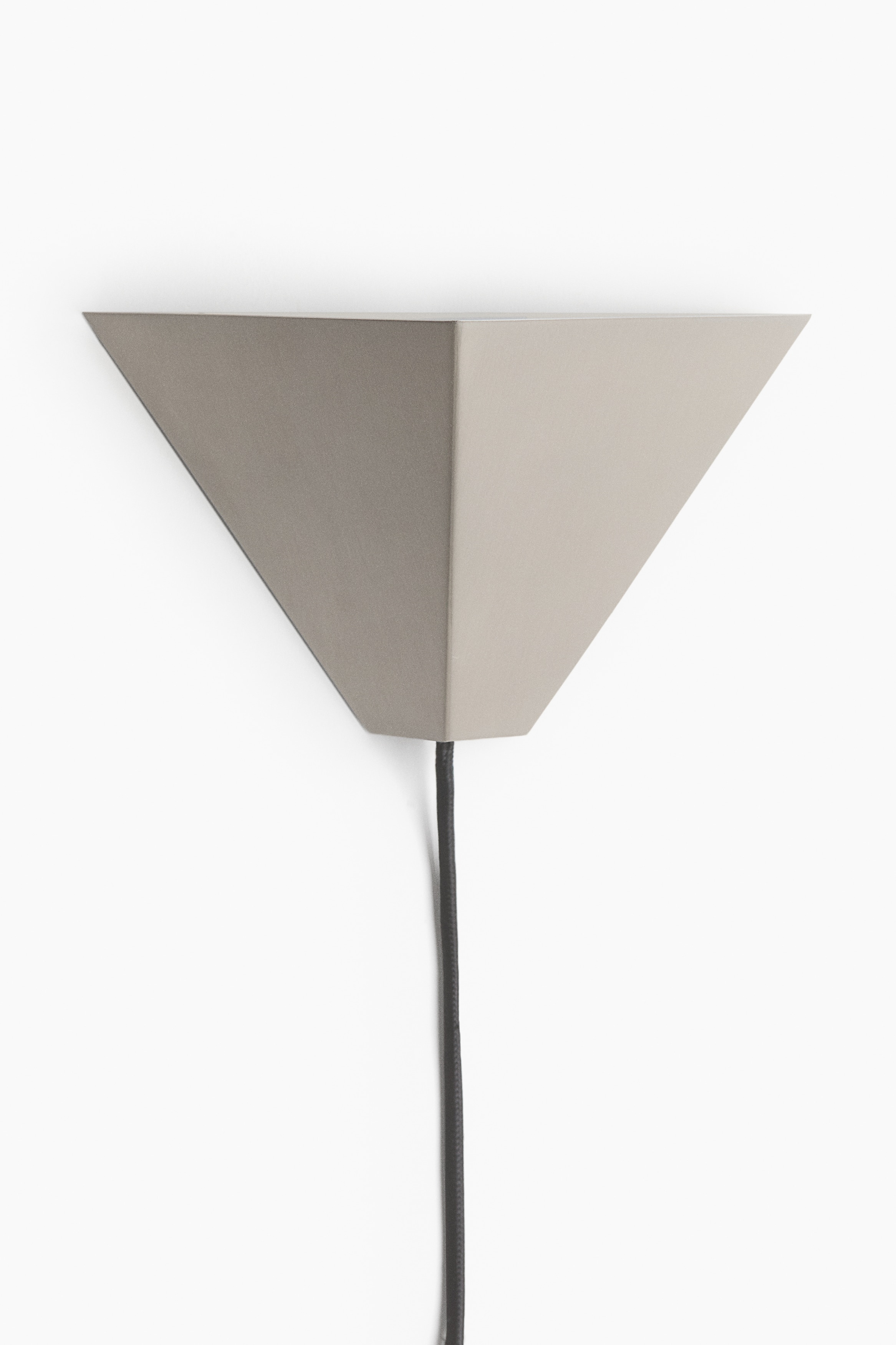 A geometric metal wall light with a dark brushed finish is shown from the front, featuring an angular inverted triangular prism shape with a central vertical crease. The lamp is illuminated, casting a warm, upward-directed glow, with a slim black fabric cord hanging straight down from its base.