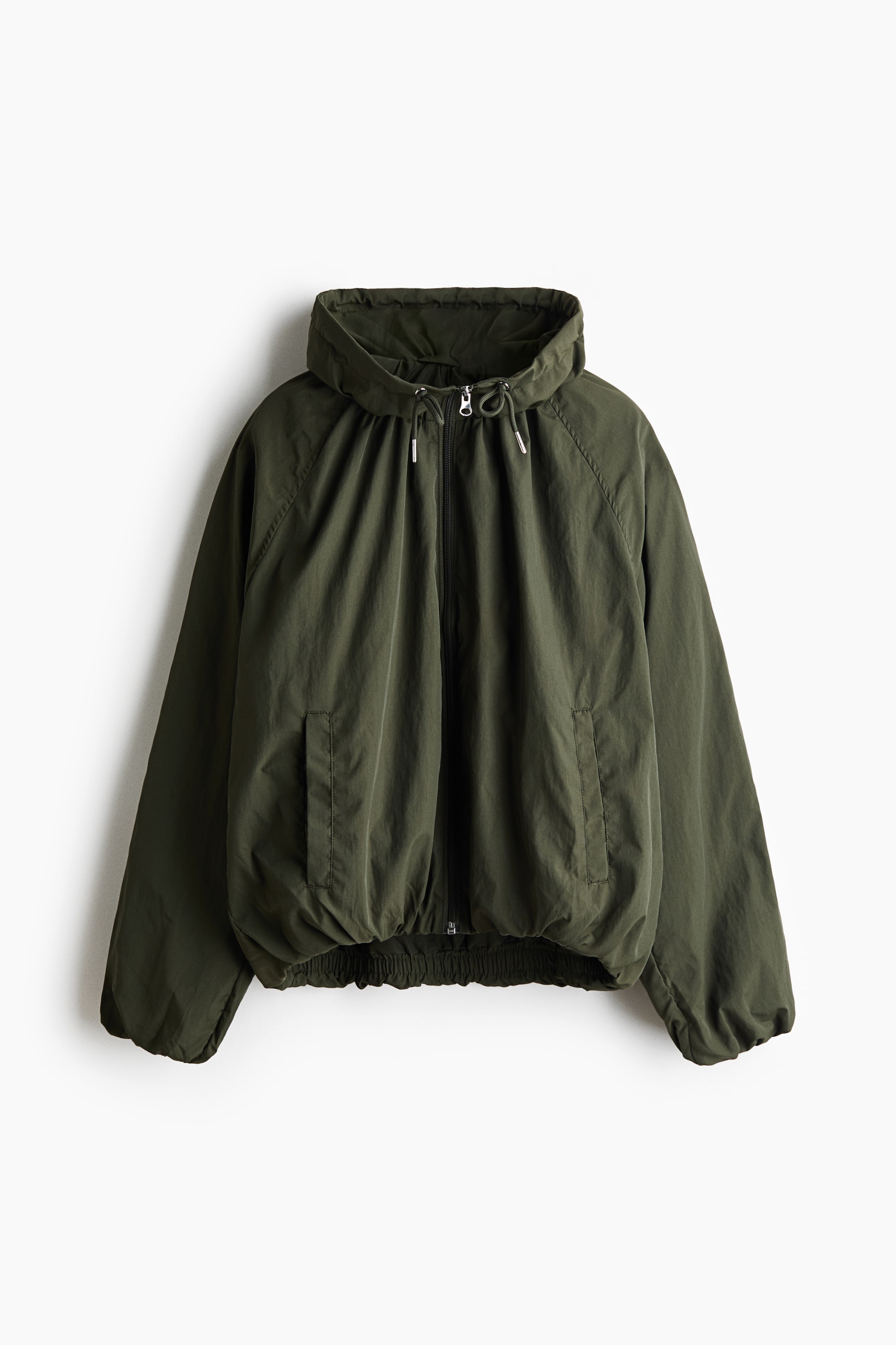 Ver imagem maior: É apresentada de frente uma jaqueta curta e oversized em nylon verde-escuro, com gola alta tipo funil com cordão ajustável e fecho de correr frontal integral. O modelo apresenta mangas raglã com punhos elásticos, bolsos embutidos verticais e uma bainha balão elástica que cria uma silhueta volumosa.
