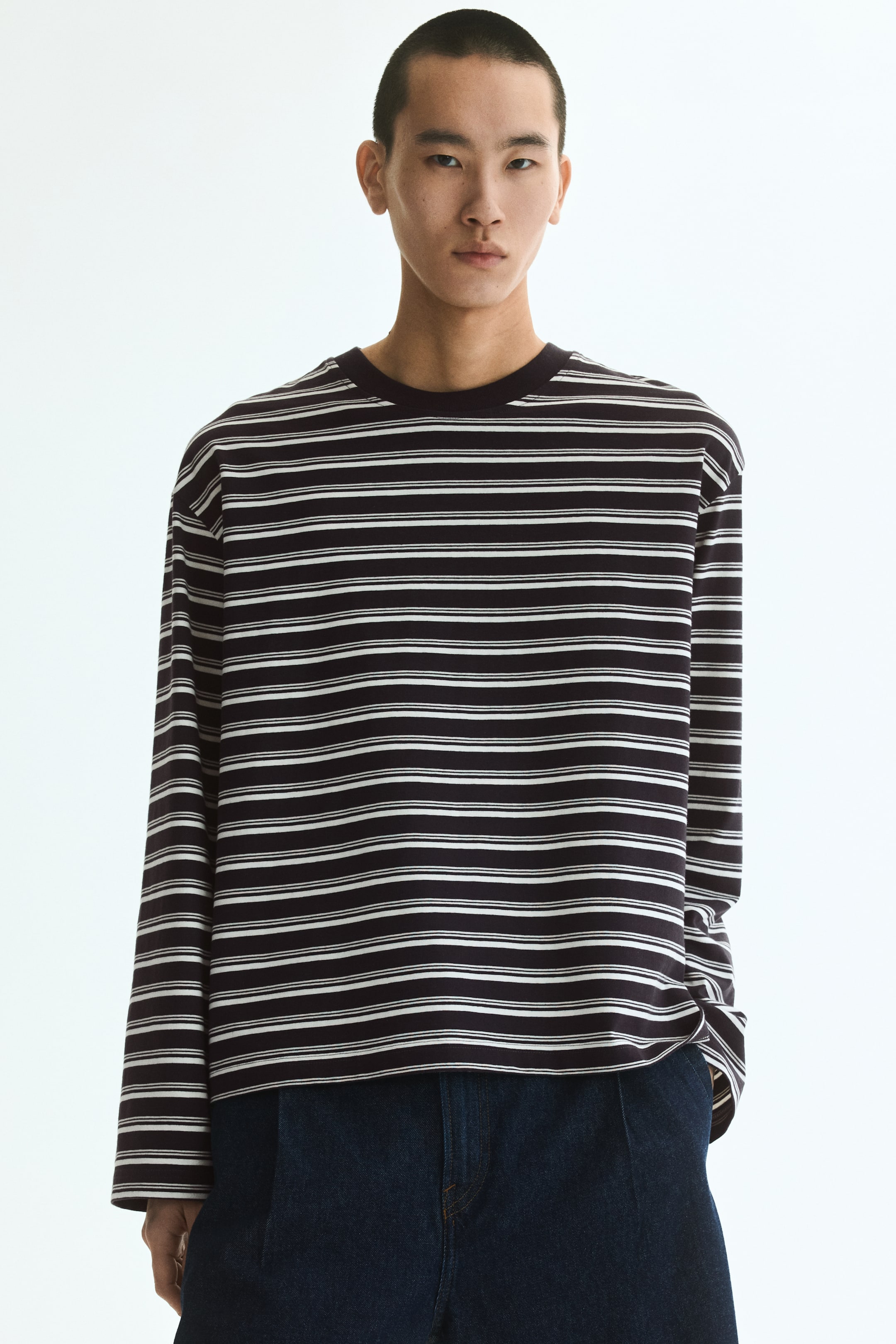 Grotere afbeelding bekijken: Een lang mouw t-shirt wordt van voren gepresenteerd, met een donkere basis en dunne witte horizontale strepen. Het heeft een ronde halslijn met ribboord, verlaagde schouders en een recht gesneden zoom, wat een ontspannen, boxy pasvorm toont.