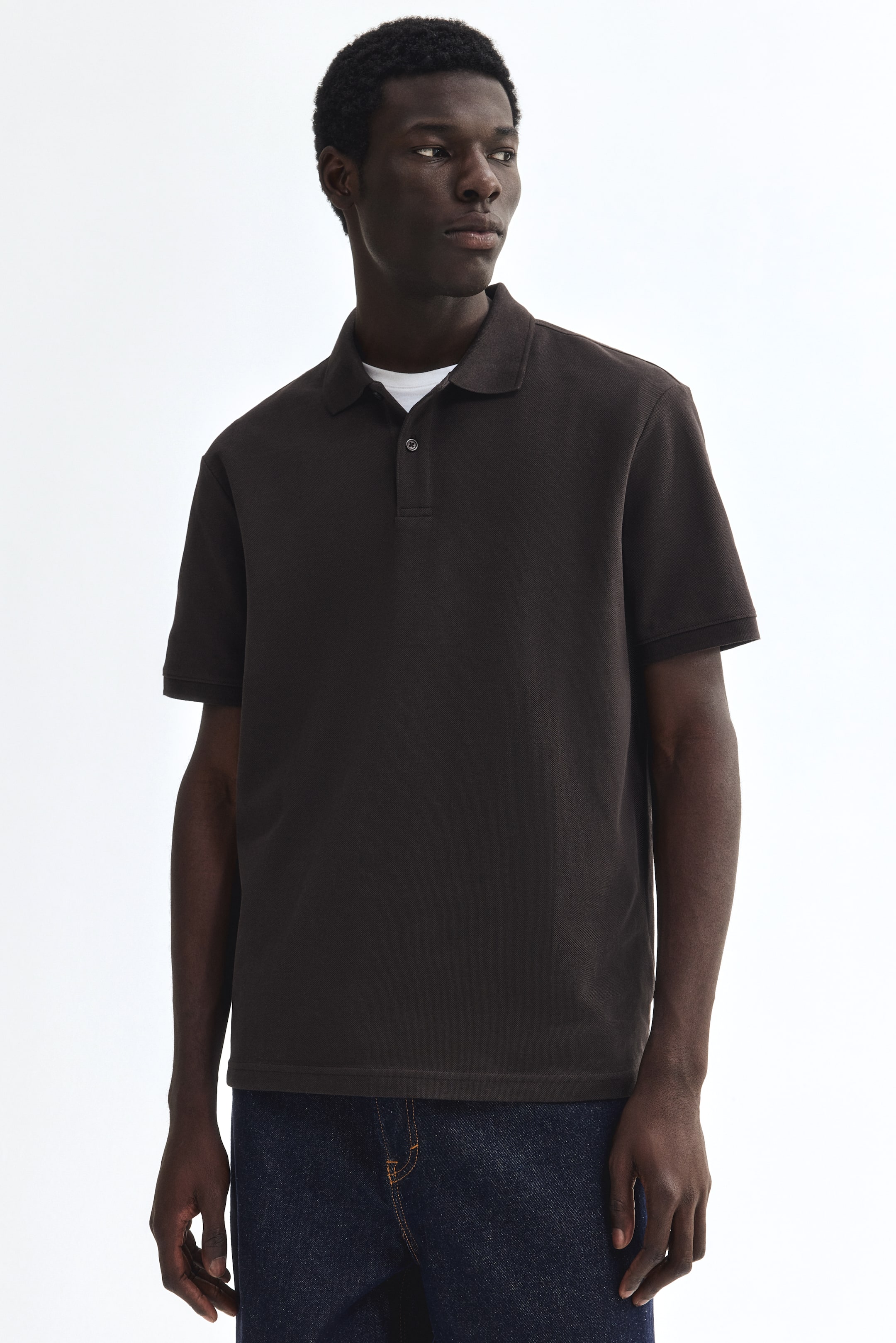Visualizza l'immagine ingrandita: Una polo in maglia marrone scuro presenta un colletto classico e una chiusura a due bottoni. È caratterizzata da maniche corte con polsini a coste e un orlo dritto, indossata sopra una maglietta bianca visibile.