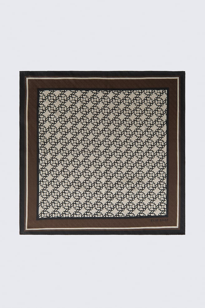 Este lenço de seda de tecelagem jacquard quadrado encontra-se estendido, exibindo um padrão entrelaçado de monogramas pretos sobre um fundo bege. A área central com padrão é emoldurada por uma espessa borda castanha com uma fina faixa creme e um rebordo exterior preto, com o nome da marca "ELIE SAAB" impresso no canto inferior direito. - 1