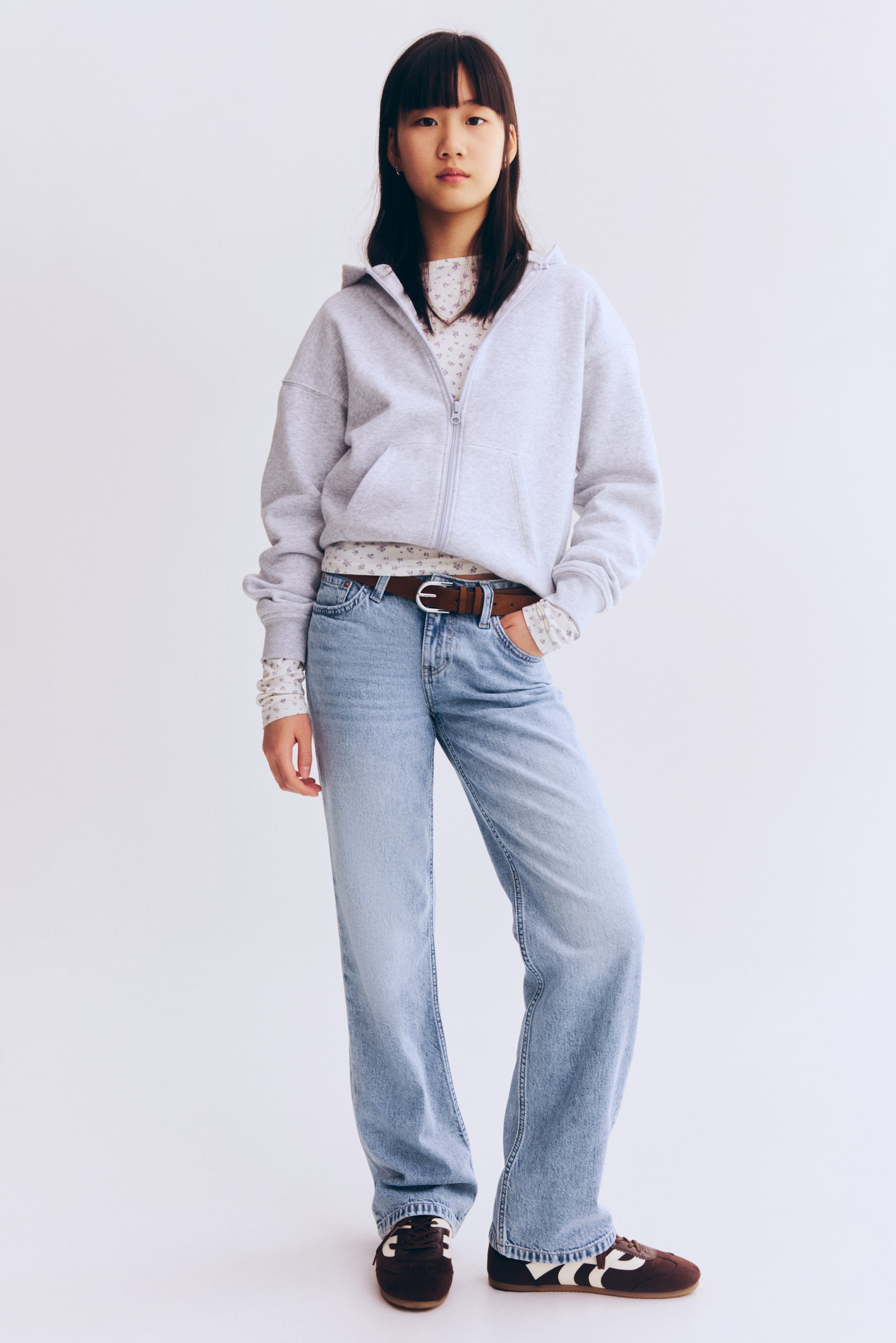Größeres Bild ansehen: Ein hellgrauer Kapuzenpullover mit durchgehendem Reißverschluss wird über einem weißen, langärmeligen Oberteil mit Blumenmuster getragen und mit einer hell gewaschenen Jeans mit weitem Bein sowie einem braunen Gürtel kombiniert. Dunkelbraune Sneakers mit weißen Streifen vervollständigen das Outfit, das in einer vollständigen Frontalansicht präsentiert wird.