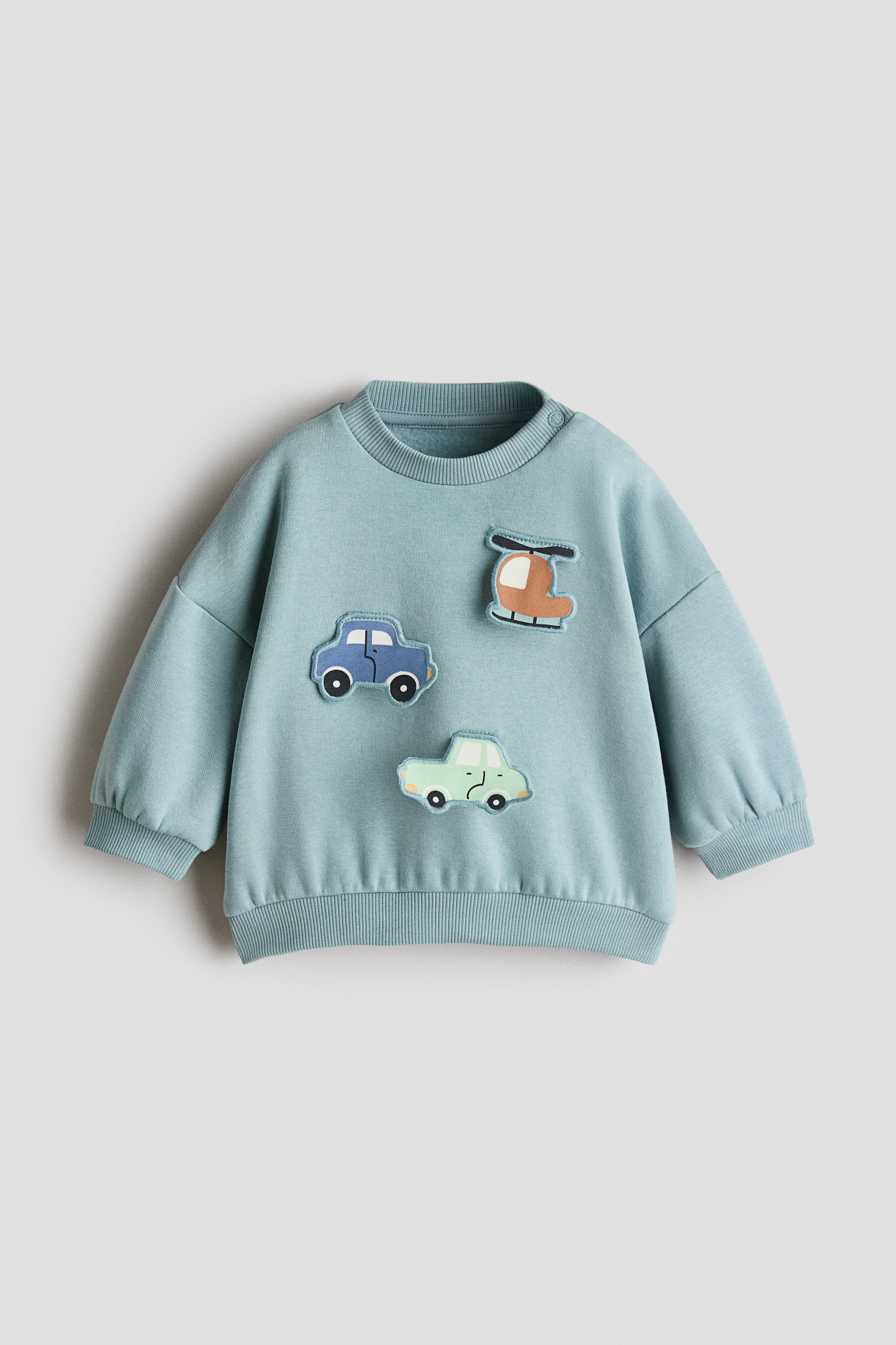 View larger image: An aqua blue sweatshirt is on display, showcasing its front with a ribbed round neck and two snap buttons on the left shoulder. It features long sleeves with ribbed cuffs, a ribbed hem, and three appliqué patches on the front: a brown helicopter, a blue car, and a light green car.