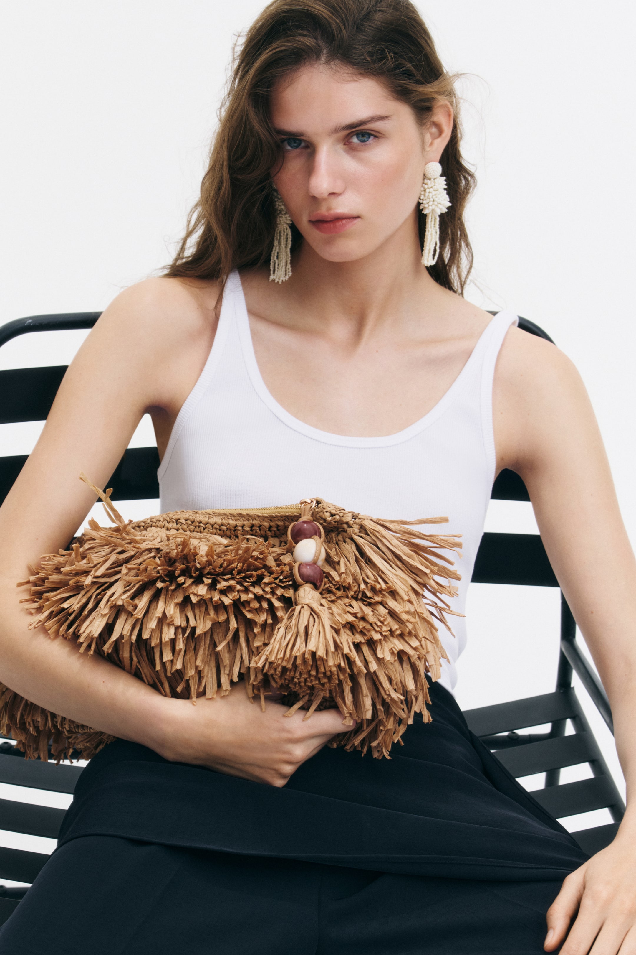 Se større billede: En frynset raffia clutch taske, der har en dekorativ kvast med to perler og en synlig lynlås, bliver holdt af modellen.