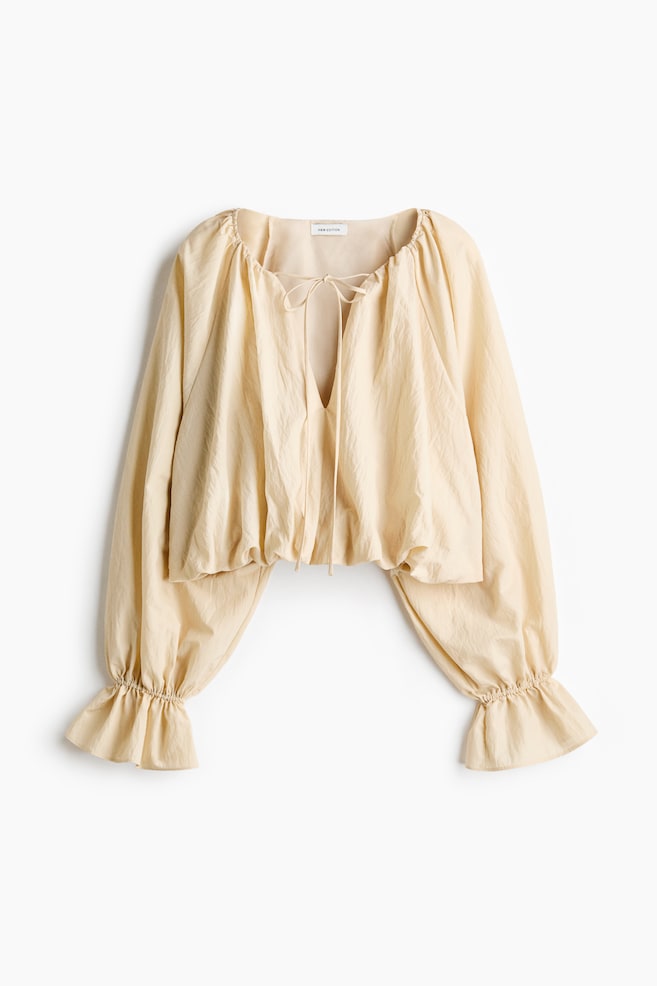A cream top is displayed from the front, showcasing a gathered round neckline with a V-cut opening and ties knotted at the top. It features long, voluminous sleeves that end in ruffled cuffs and an elasticized hem, crafting a cropped silhouette. - 1