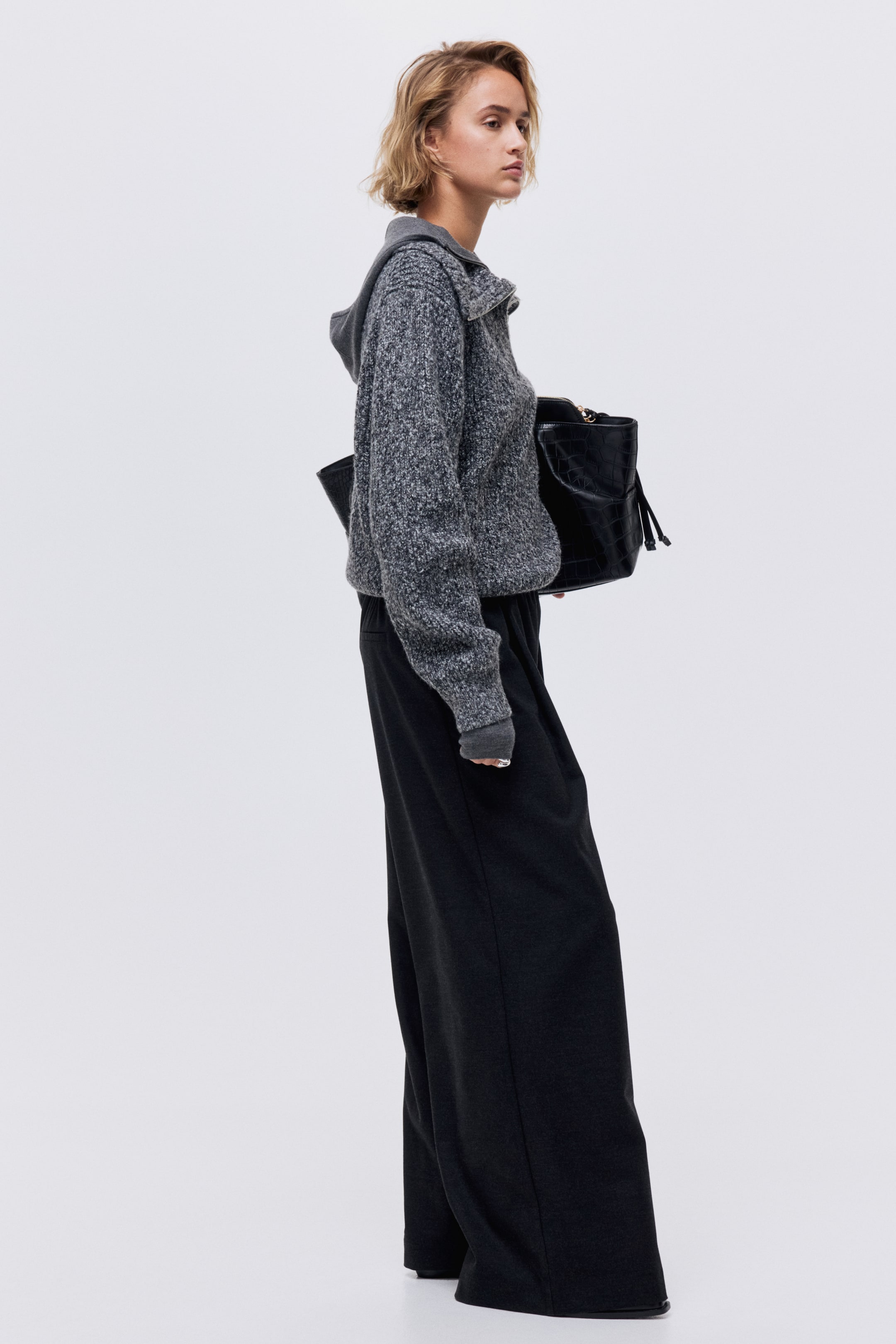 Näytä suurempi kuva: Vaaleanharmaa neulottu puolivetoketjullinen paita, jossa on vaaleanharmaa huppu ja ribatut hihansuut, esitellään sivuprofiilista yhdessä tummanharmaiden leveälahkeisten housujen kanssa.