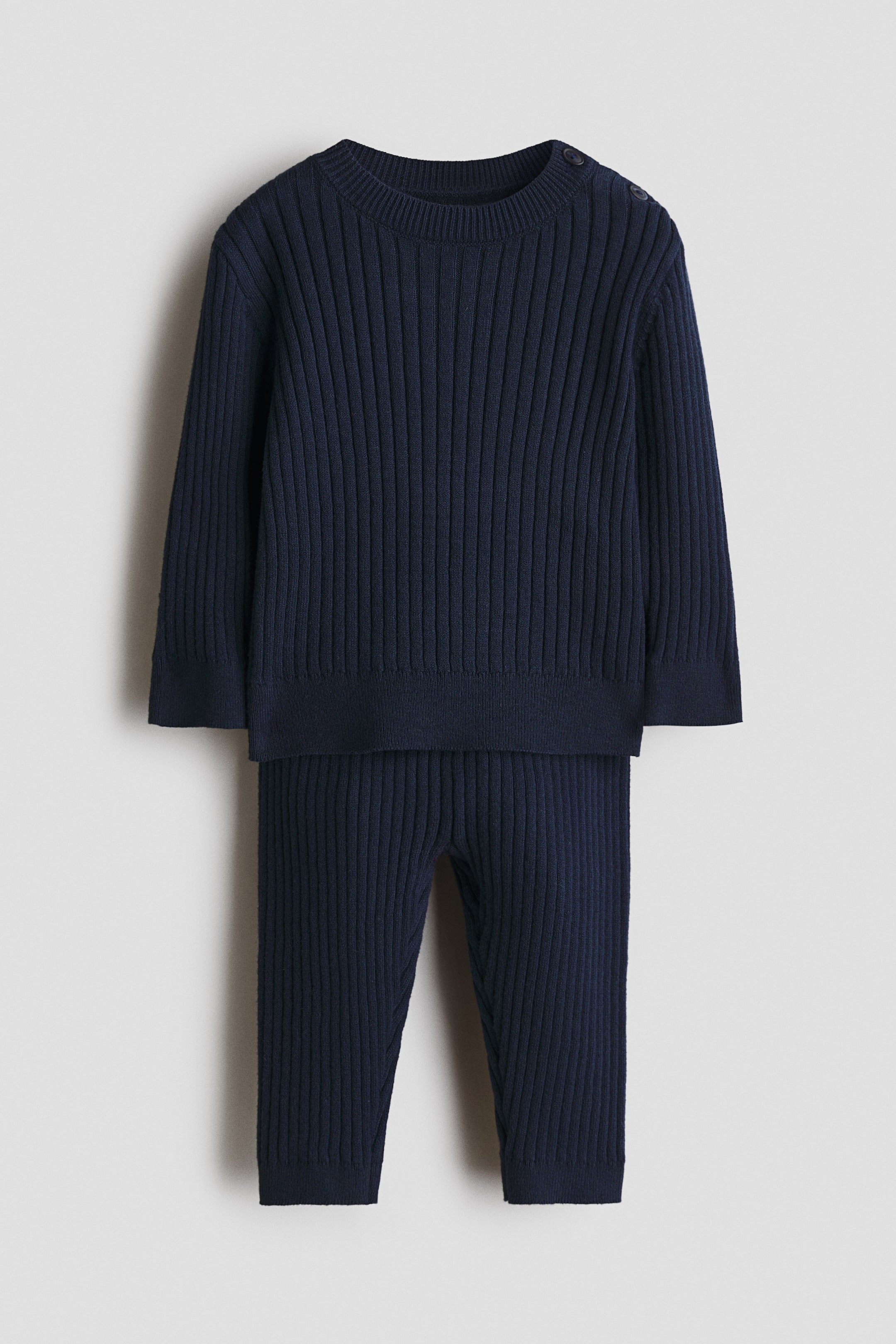 Größeres Bild anzeigen: Ein marineblaues Strick-Set mit breiter Rippstruktur wird präsentiert und besteht aus einem Pullover und einer Hose. Der frontal gezeigte Pullover verfügt über einen Rundhalsausschnitt, zwei Knöpfe an der linken Schulter sowie fein gerippte Abschlüsse an Halsausschnitt, Ärmelbündchen und Saum, während die Hose einen fein gerippten Bund und fein gerippte Beinabschlüsse zeigt.