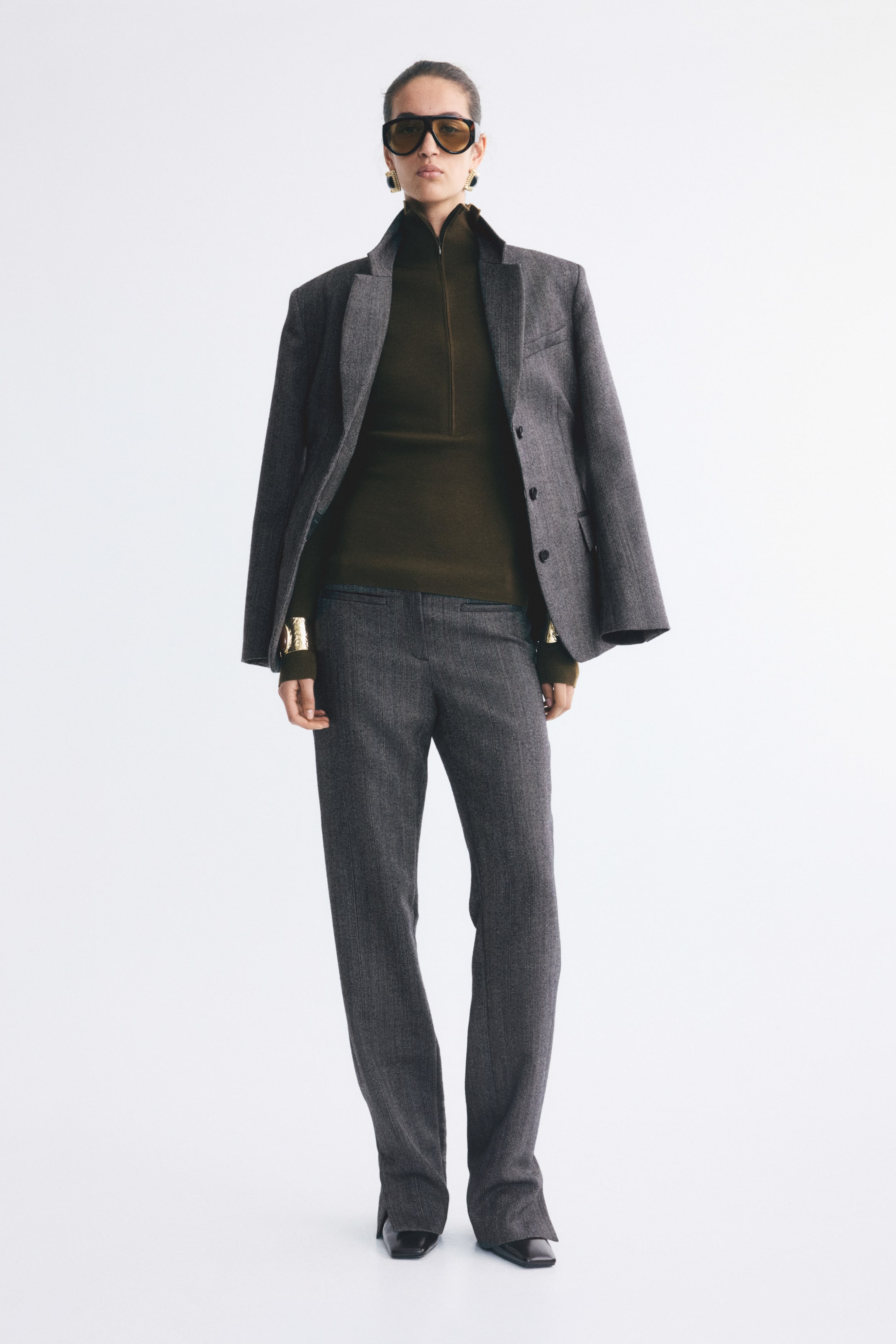 View larger image: The model is wearing trousers in a grey textured wool weave, featuring a low waist, straight legs, and high slits at the hems. The trousers are styled with a matching grey blazer draped over the shoulders and a dark green high-neck top.