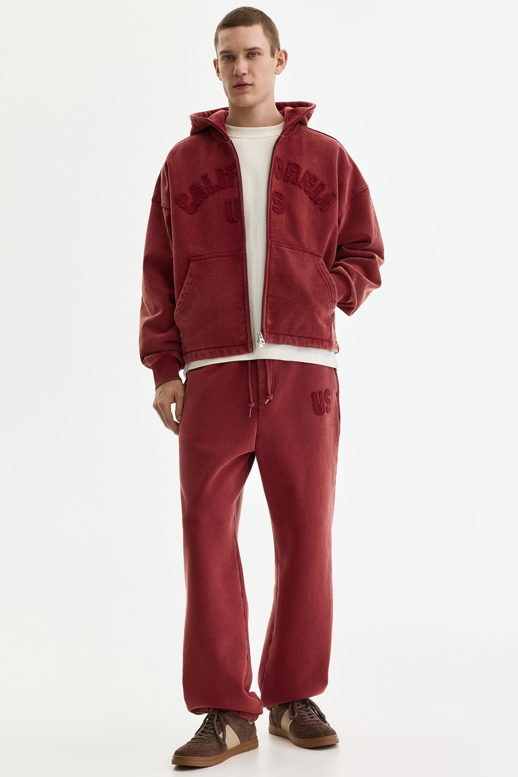 View larger image: A washed red hooded tracksuit is shown, featuring an oversized full-zip hoodie with "CALIFORNIA US" embroidered on the chest and large front pockets. The hoodie is worn over a cream t-shirt, complementing matching wide-leg sweatpants that gather at the ankles and have "US" embroidered on the left thigh.