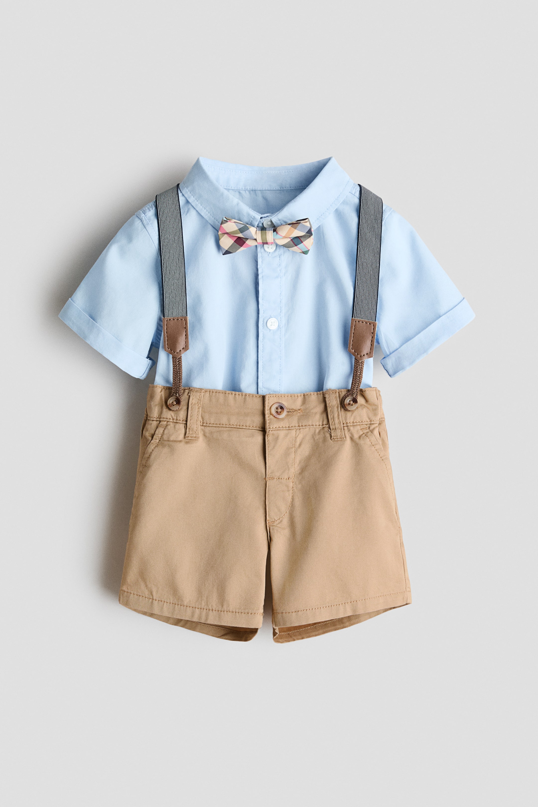 Ingrandisci l'immagine: Un completo elegante presenta una camicia azzurra a maniche corte con colletto abbottonato e maniche arrotolate, indossata con pantaloncini kaki, tutti rivolti in avanti. Un papillon a quadri è coordinato con la camicia, e bretelle a righe scure con linguette marroni sono attaccate ai pantaloncini, che presentano tasche laterali e una patta finta con bottone.