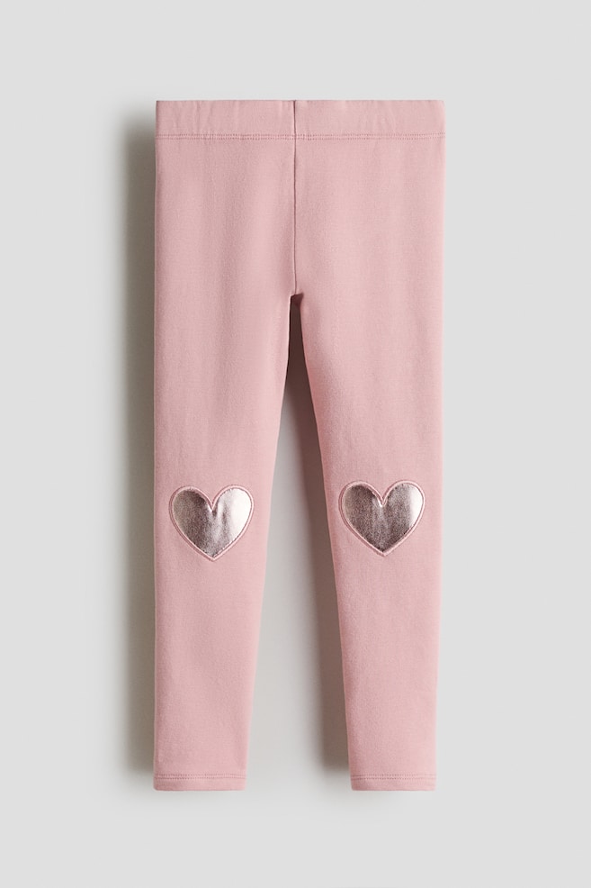 Front-facing pink leggings are displaying a soft elasticated waistband and two metallic, rose gold-toned heart patches on the knees. - 1