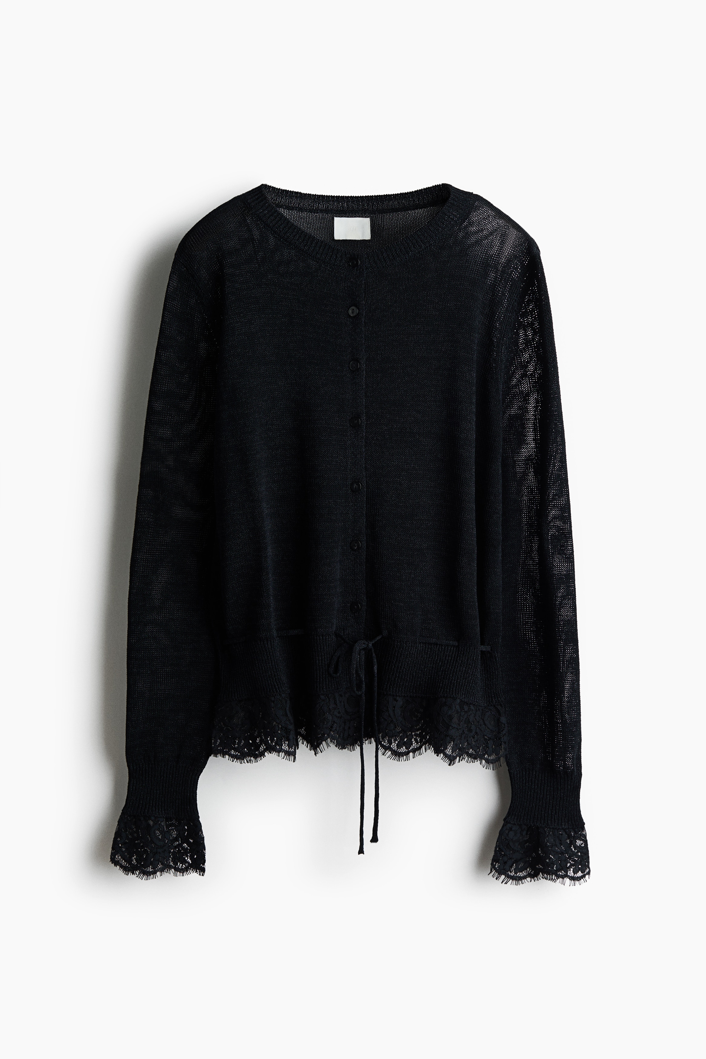A dark, fine-knit cardigan featuring a buttoned front, long sleeves with lace-trimmed cuffs, and a lace-trimmed hem is being worn by the model, who is standing with her arms crossed. The cardigan is paired with matching dark trousers and pointed-toe shoes.