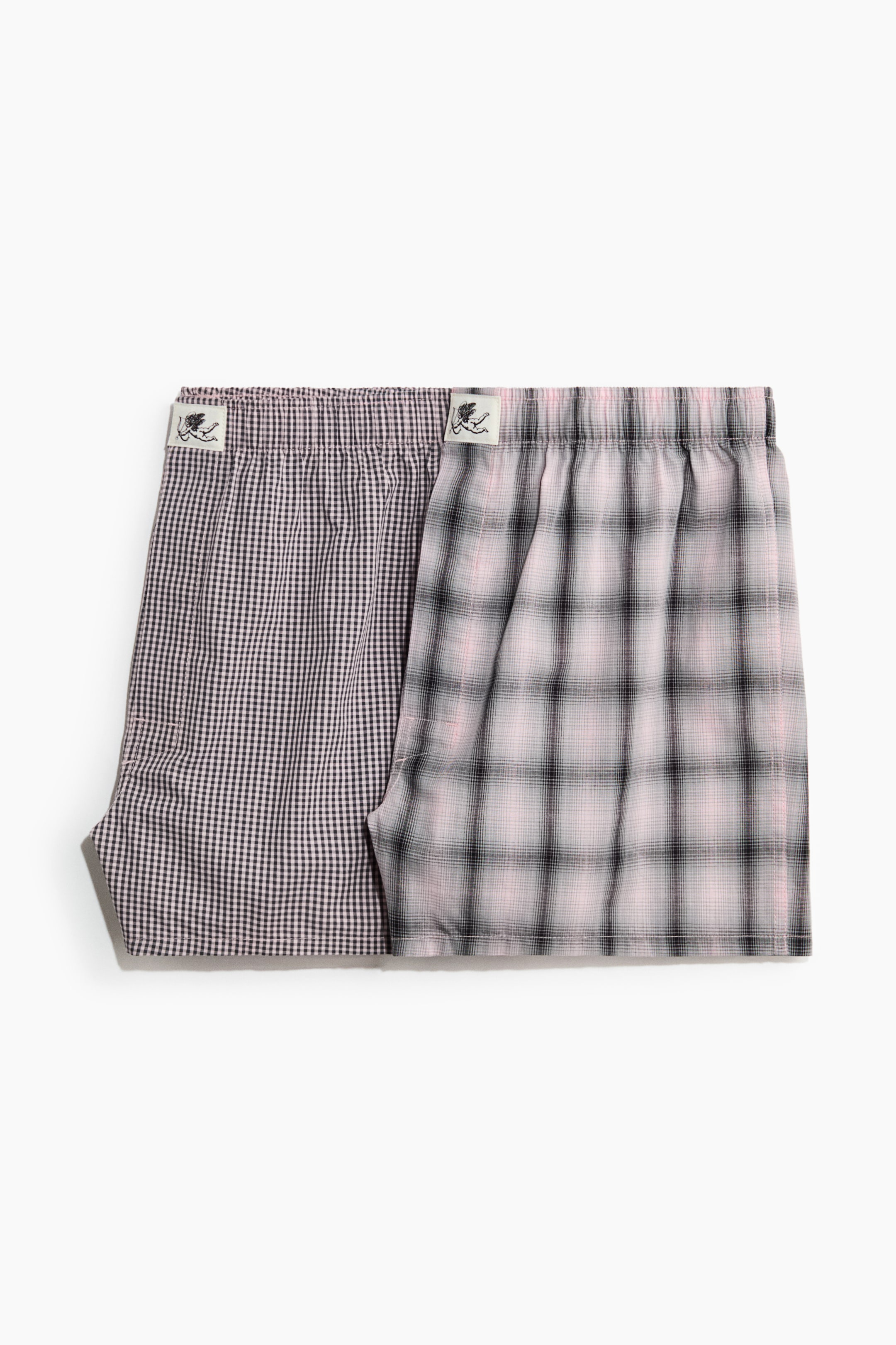 View larger image: Two pairs of boxer shorts are lying side-by-side, presenting their fronts; the left pair is featuring a black and white gingham check pattern, and the right pair is displaying a pink and black plaid pattern. Both shorts are designed with an elasticated waist, each accented by a small graphic tag.