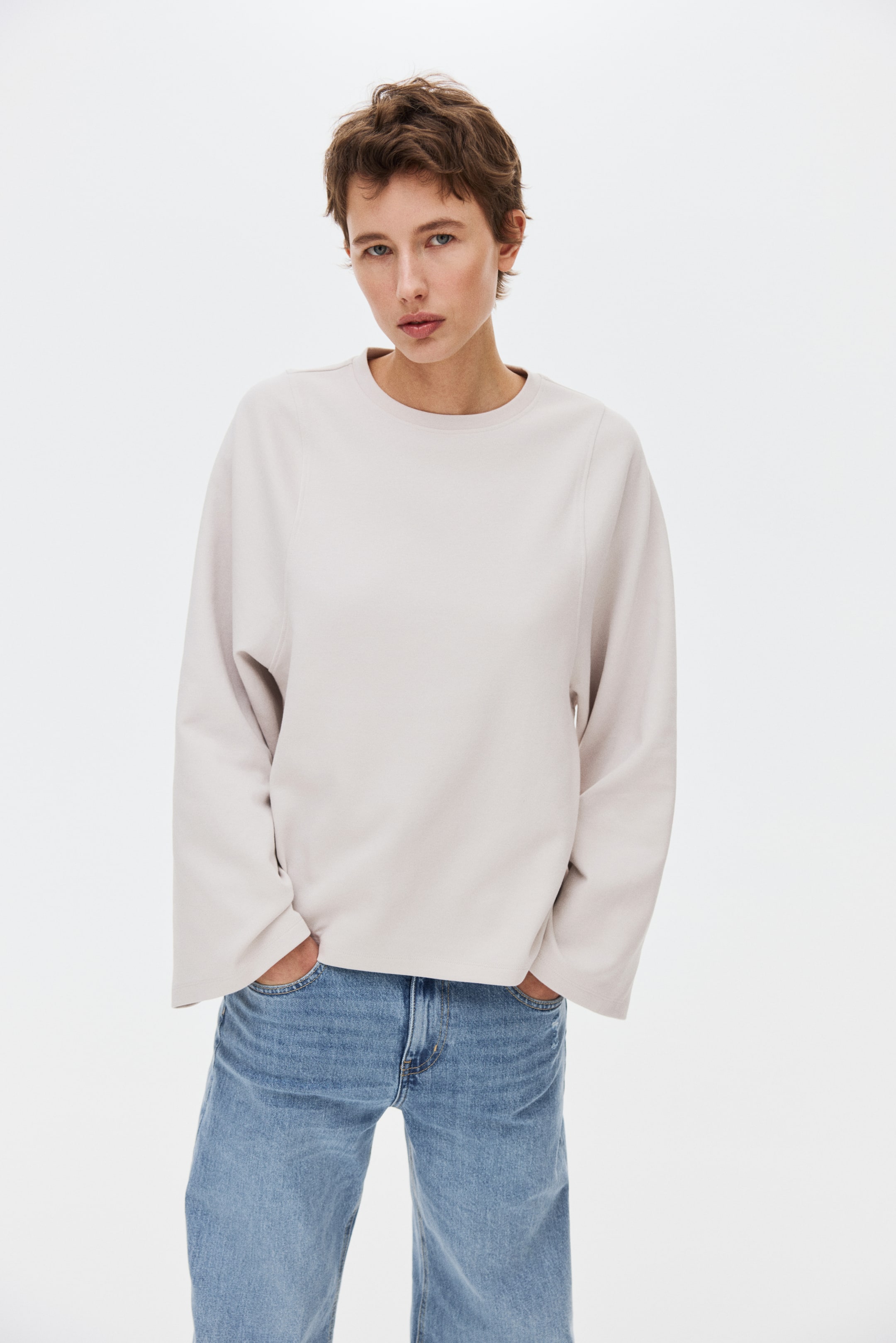 View larger image: A light beige crew-neck top is cut in a relaxed, slightly oversized fit. It features long, wide sleeves with dropped shoulders and distinctive seam detailing that extends from the neckline.