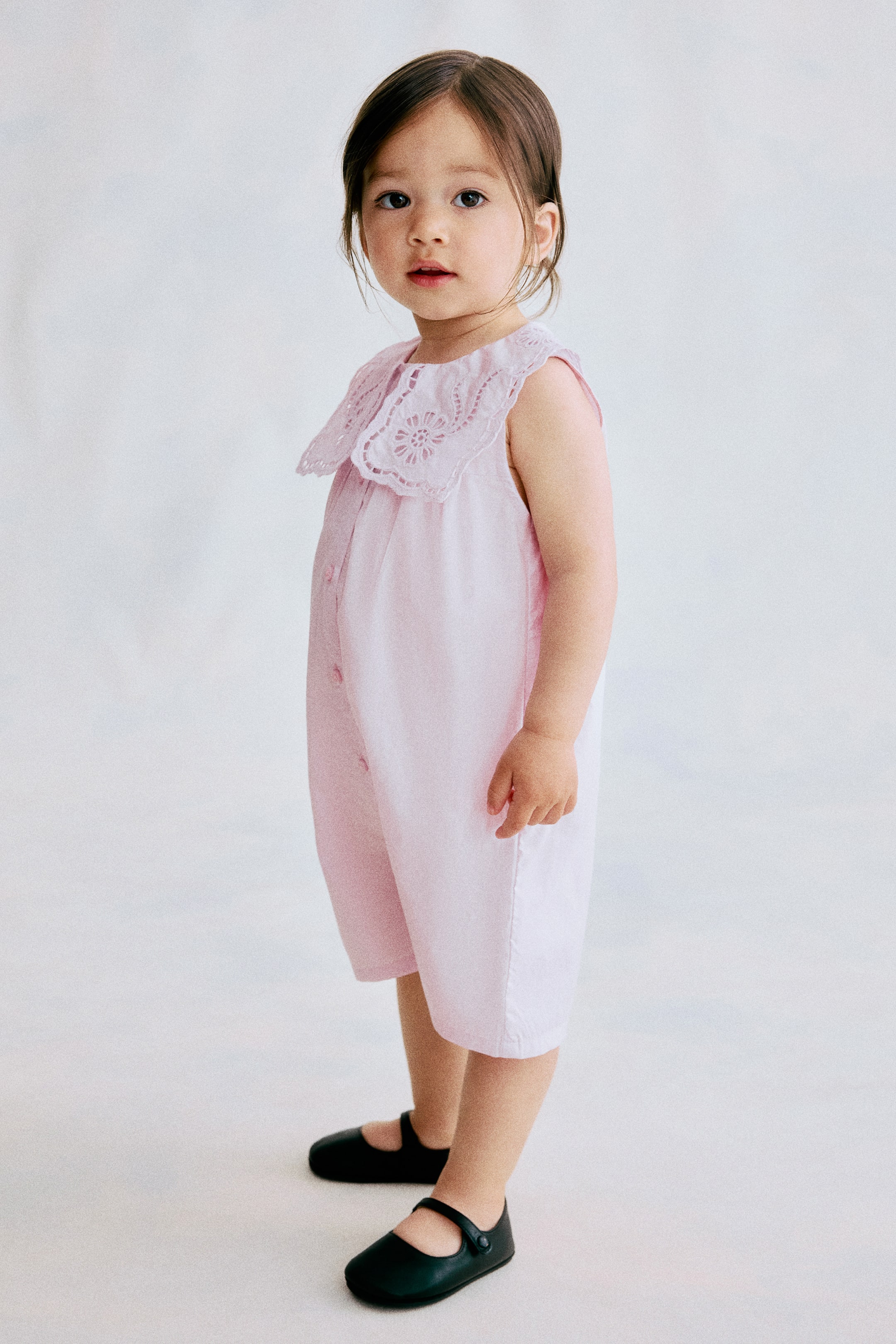 View larger image: The light pink sleeveless baby romper is shown from a slight angle, featuring a large, round, eyelet embroidered collar. A line of buttons is running down the front, and its short legs are extending above the knees.