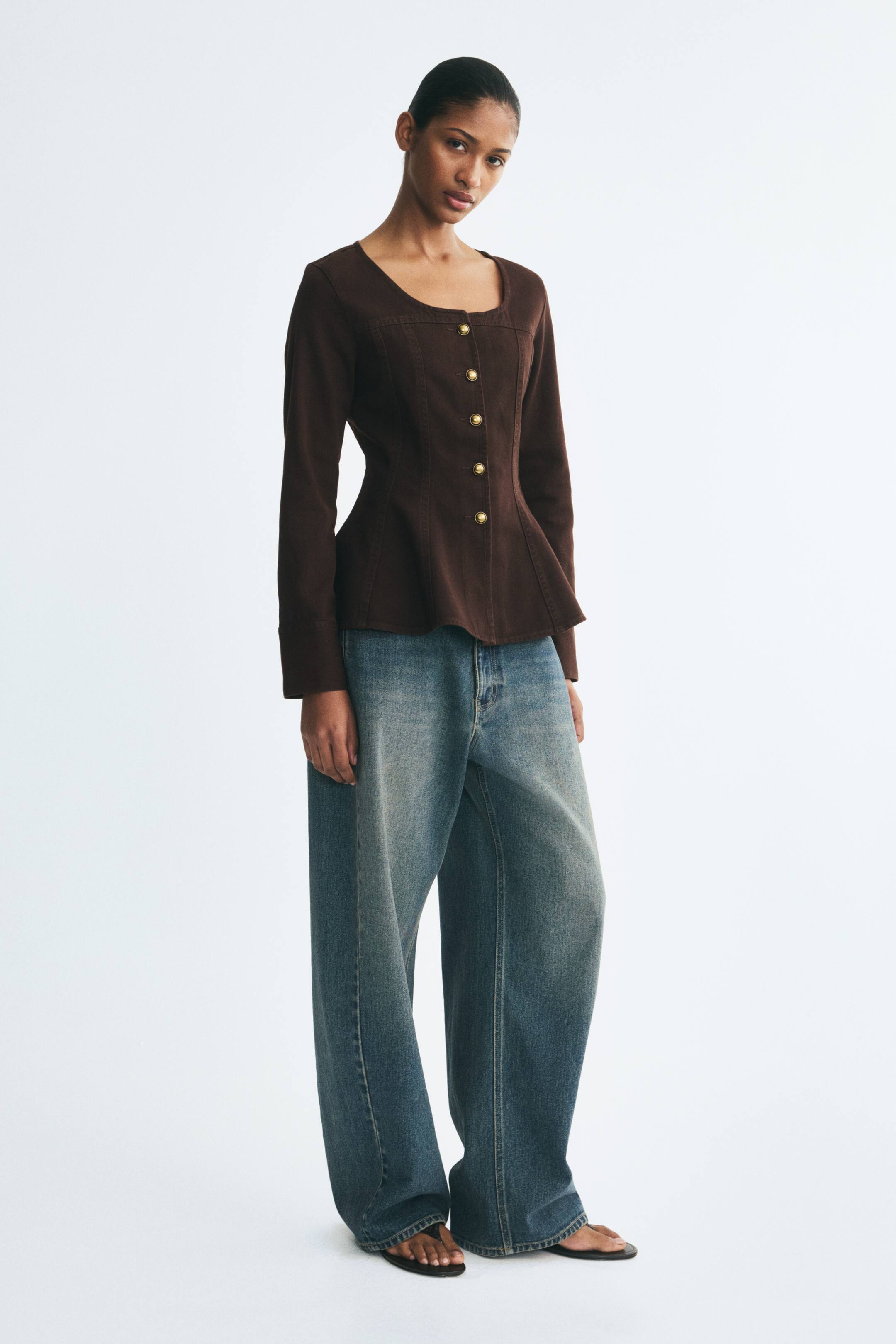 View larger image: The model is wearing a fitted dark brown denim jacket featuring a low round neckline, gold metal buttons down the front, visible vertical seams, and a flared peplum hem. The jacket is styled with wide-leg blue jeans.