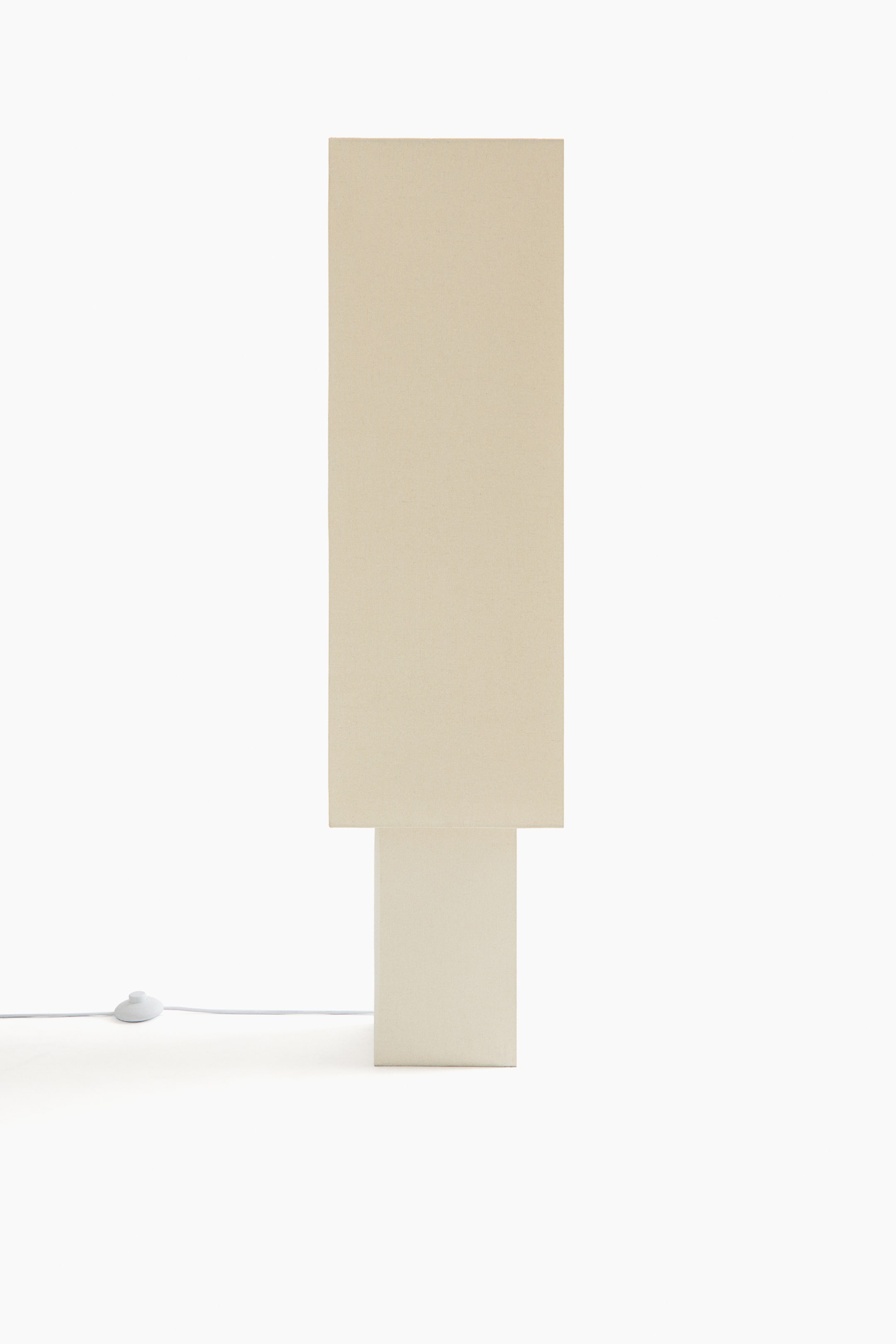 View larger image: A beige pillar lamp is facing front, showcasing a tall, rectangular shade resting on a shorter, wider rectangular base. A white power cord with an inline switch extends from its lower left side.