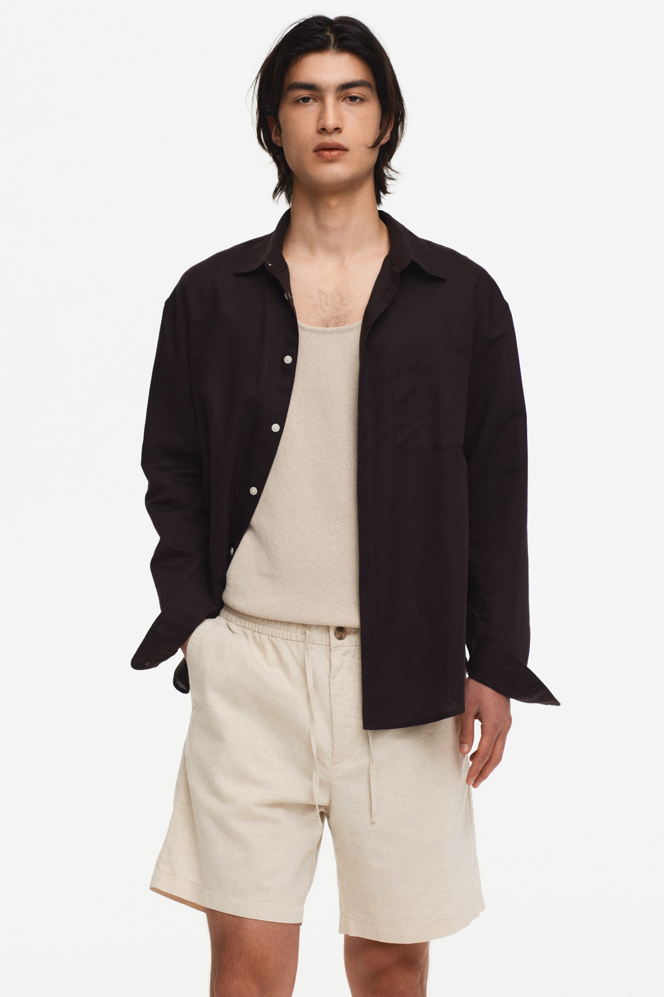 View larger image: Dark brown long-sleeve shirt featuring a spread collar, button-front closure with white buttons, and a single patch pocket on the left chest. Worn open over a light beige top, it highlights cuffed long sleeves and a relaxed fit.
