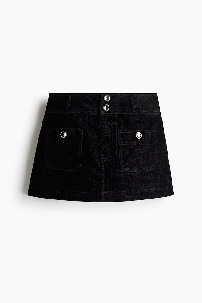 A black mini skirt is presented FRONT, featuring a low waist and constructed from soft denim. It is designed with a zip fly, two rhinestone buttons at the waistband, and two large front patch pockets, each adorned with a single rhinestone button. - 1