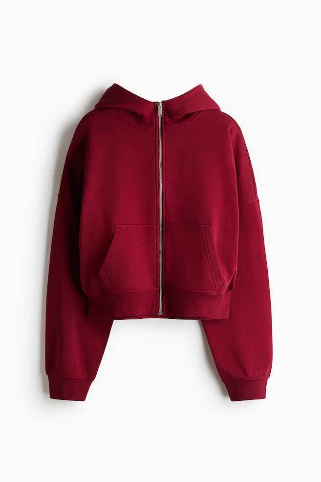 Ein locker sitzender roter Hoodie aus Sweatshirtstoff wird von vorne gezeigt, mit einer gefütterten Kapuze, einem silbernen Reißverschluss vorne und aufgesetzten Vordertaschen. Er verfügt außerdem über überschnittene Schultern, lange Ärmel mit gerippten Bündchen und einen gerippten Saum. - 1