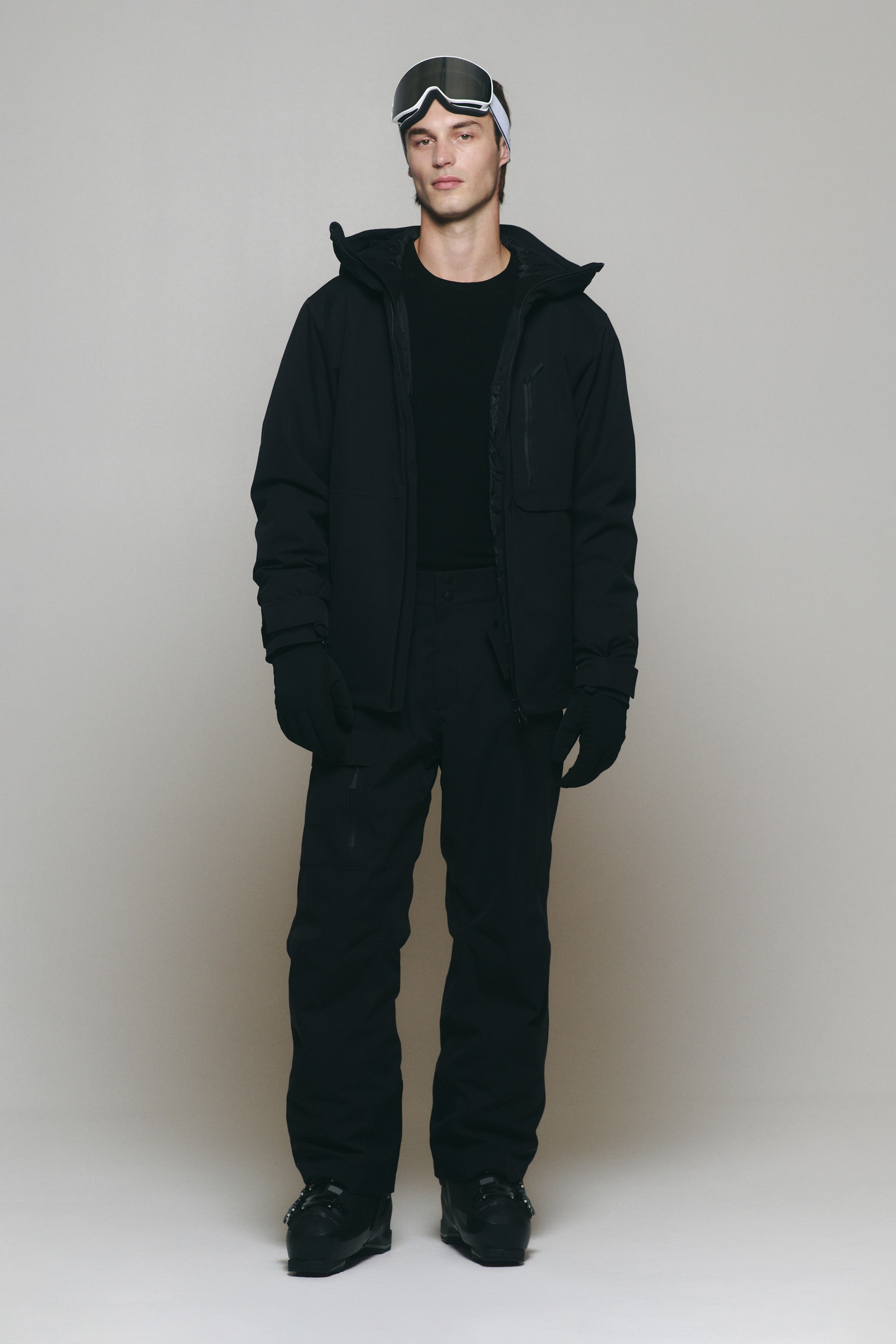 Größeres Bild ansehen: Das Model trägt eine schwarze Skijacke mit Kapuze und eine dazu passende schwarze Skihose. Die offene Jacke wird über einem schwarzen Rundhalsoberteil getragen und mit schwarzen Handschuhen, schwarzen Skistiefeln und einer auf der Stirn getragenen Skibrille mit weißem Rahmen kombiniert.