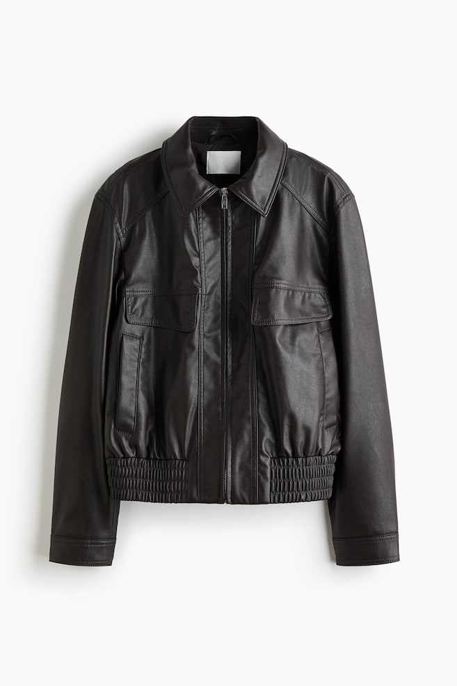 Eine dunkelbraune Lederjacke wird von vorne präsentiert, mit einem Umlegekragen und einem silberfarbenen Reißverschluss, der mittig vorne verläuft. Zwei große Pattentaschen sind im unteren Vorderbereich sichtbar, und die Jacke wird durch einen breiten, gesmokten elastischen Bund abgeschlossen. - 1