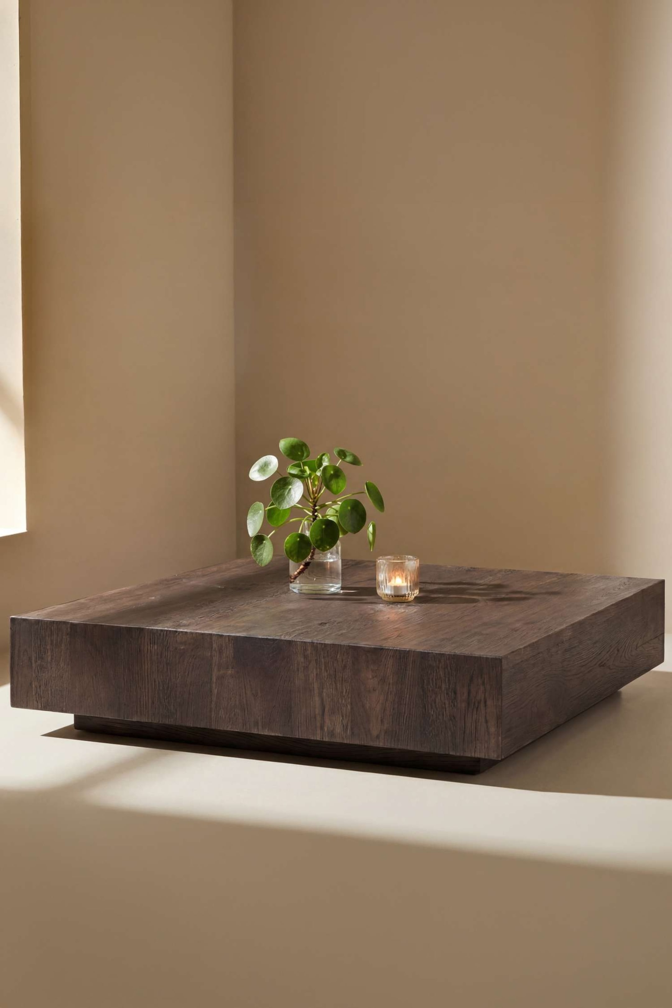 Agrandir l'image: Une table basse carrée en bois foncé présente un plateau épais avec un grain du bois visible, et repose sur un socle encastré à profil bas. Une petite plante dans un vase en verre transparent et une bougie allumée dans un photophore en verre texturé sont posées sur sa surface.
