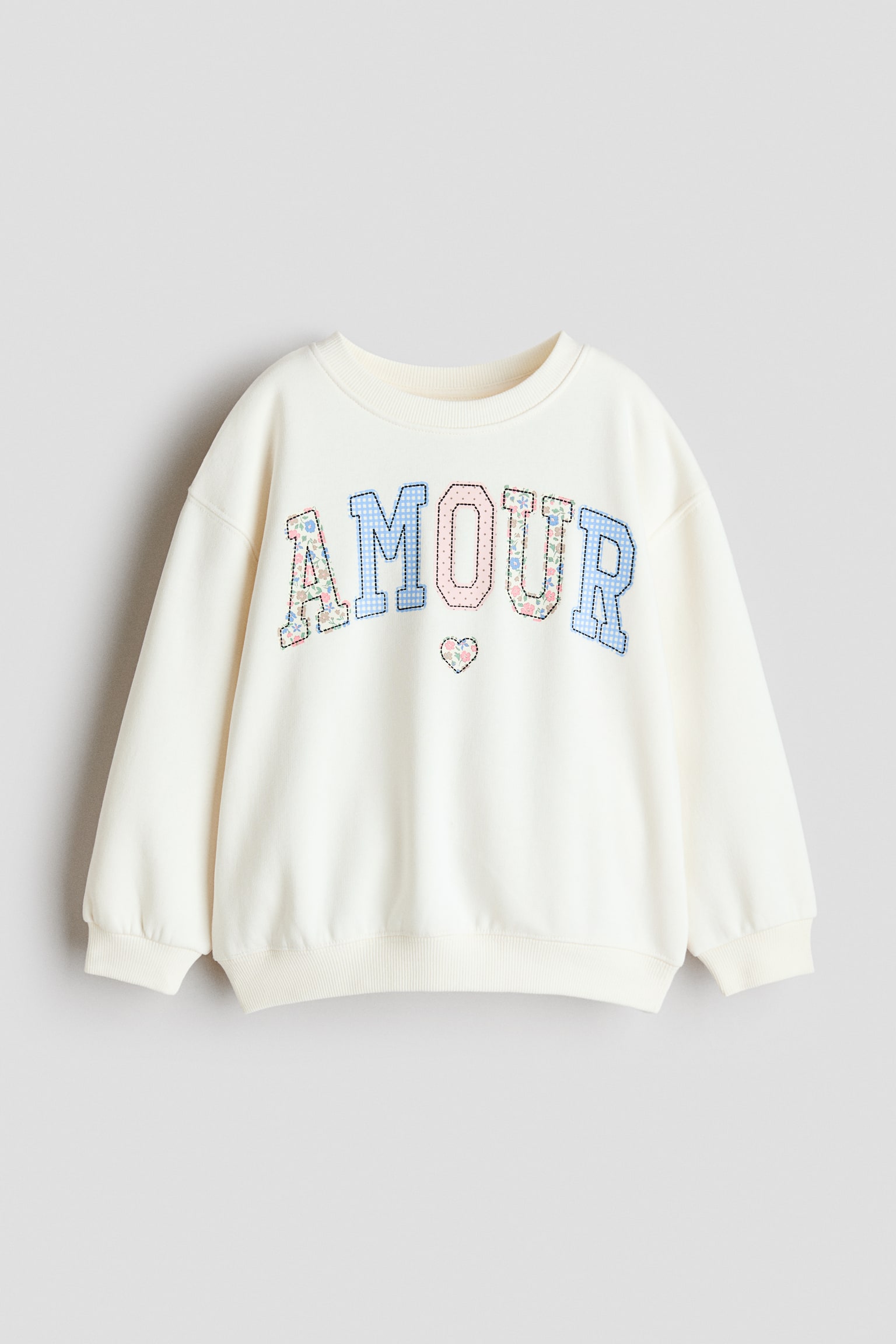 A front-facing cream sweatshirt features a ribbed crew neck, dropped shoulders, and ribbed cuffs and hem. The chest displays the word "AMOUR" in patterned appliquÃ© letters with stitched outlines, including floral and gingham designs, with a small floral heart appliquÃ© positioned below.