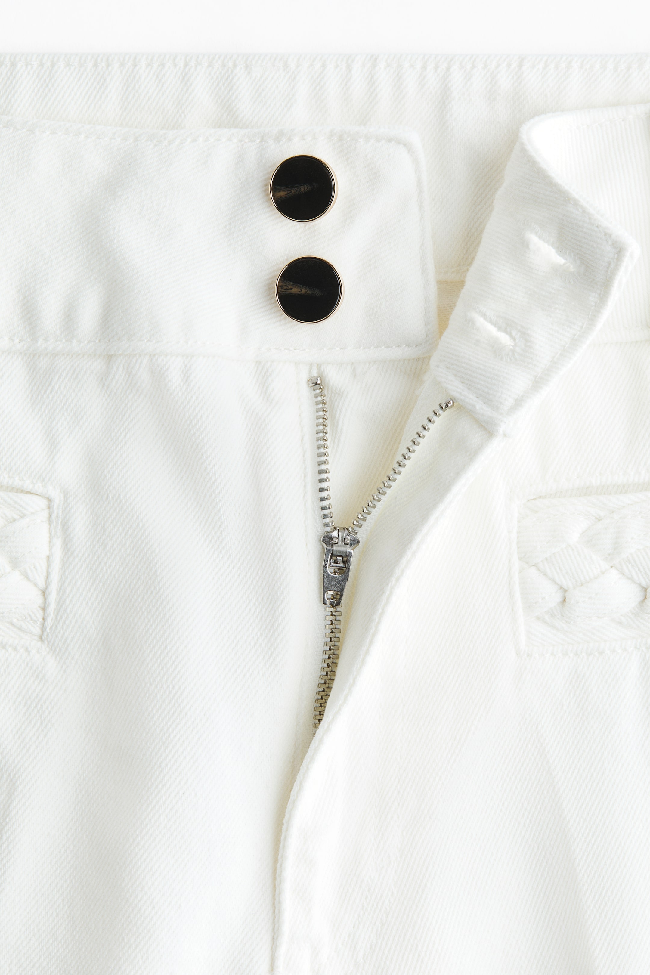 Voir une image plus grande: Le DEVANT des jeans blancs présente une braguette avec fermeture éclair partiellement déboutonnée et deux boutons ronds noirs et dorés qui sécurisent la taille haute. Un passepoil tressé est visible sur les poches avant à la française.