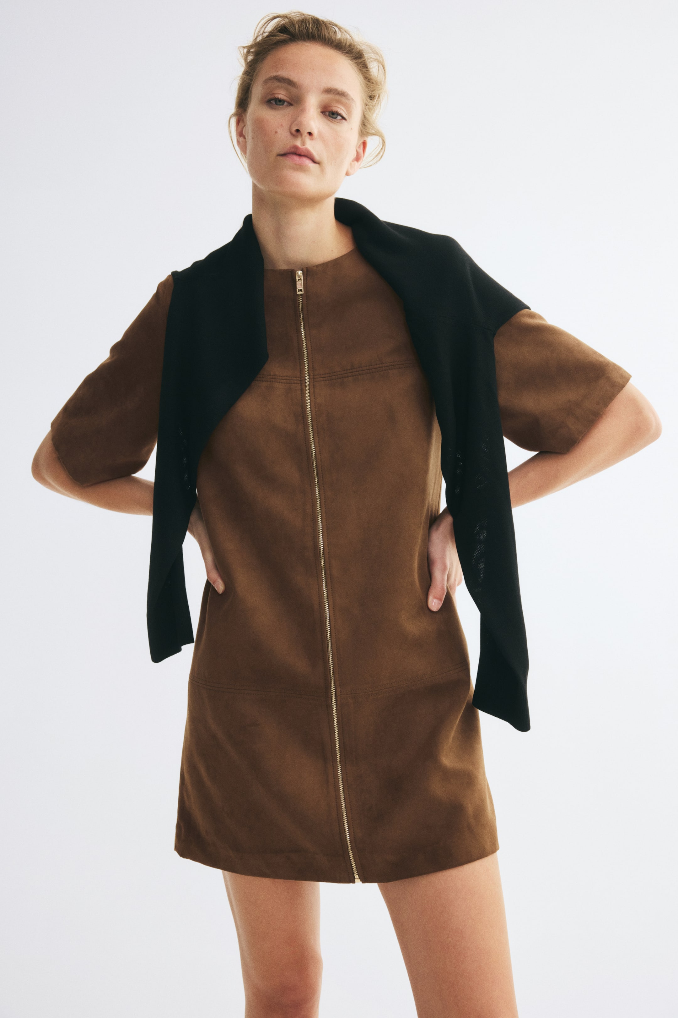 View larger image: A short brown A-line dress with a soft, brushed finish is shown from the front, featuring a round neckline, short sleeves, and a full-length gold-tone zip running down the centre. A black sweater is draped over the shoulders, framing the upper portion of the dress.