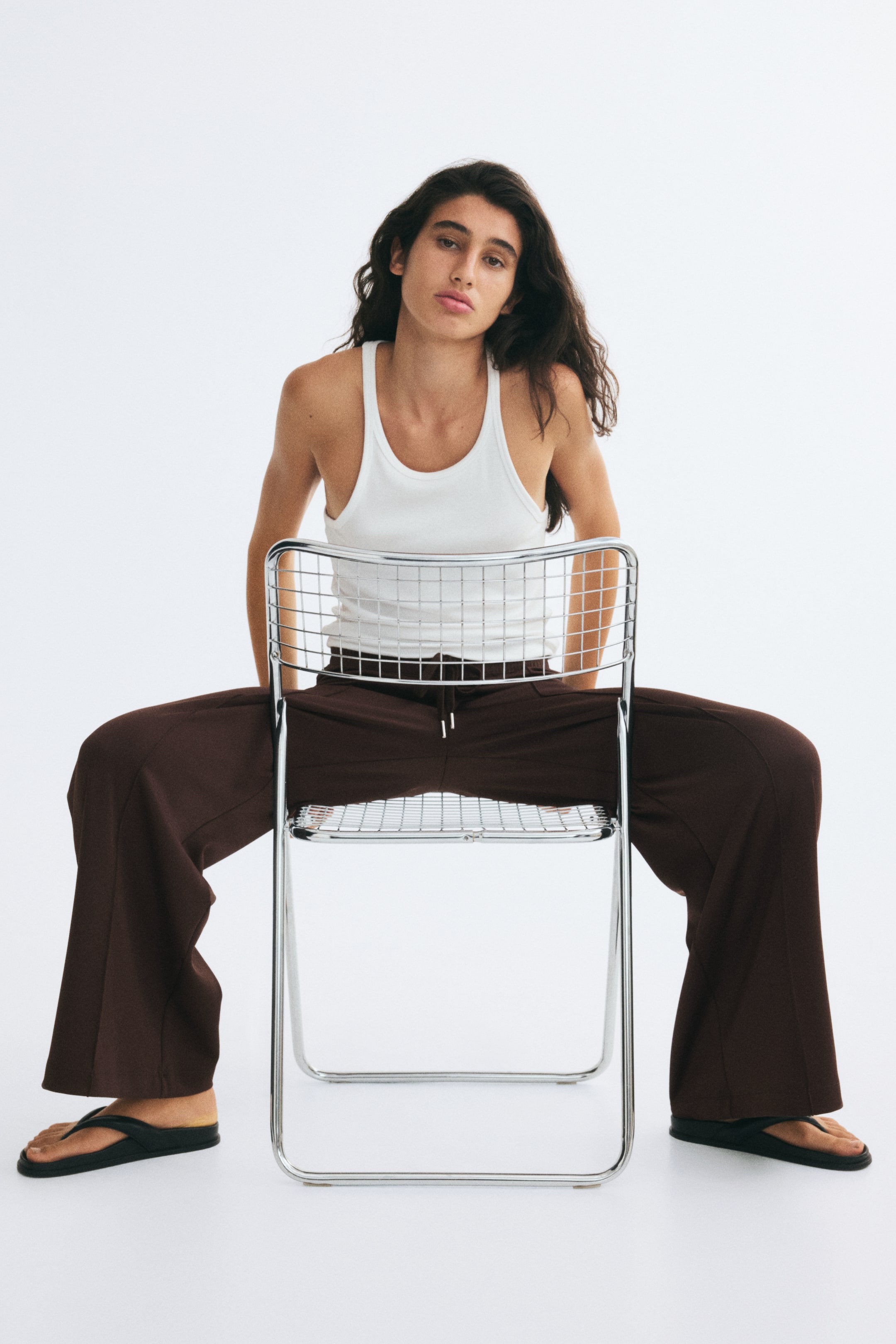 View larger image: Dark brown oversized pants featuring a high, elasticated drawstring waist and wide legs with sewn-in creases are worn by the model while sitting on a metal wire chair. The pants are styled with a tucked-in white tank top and paired with black sandals.