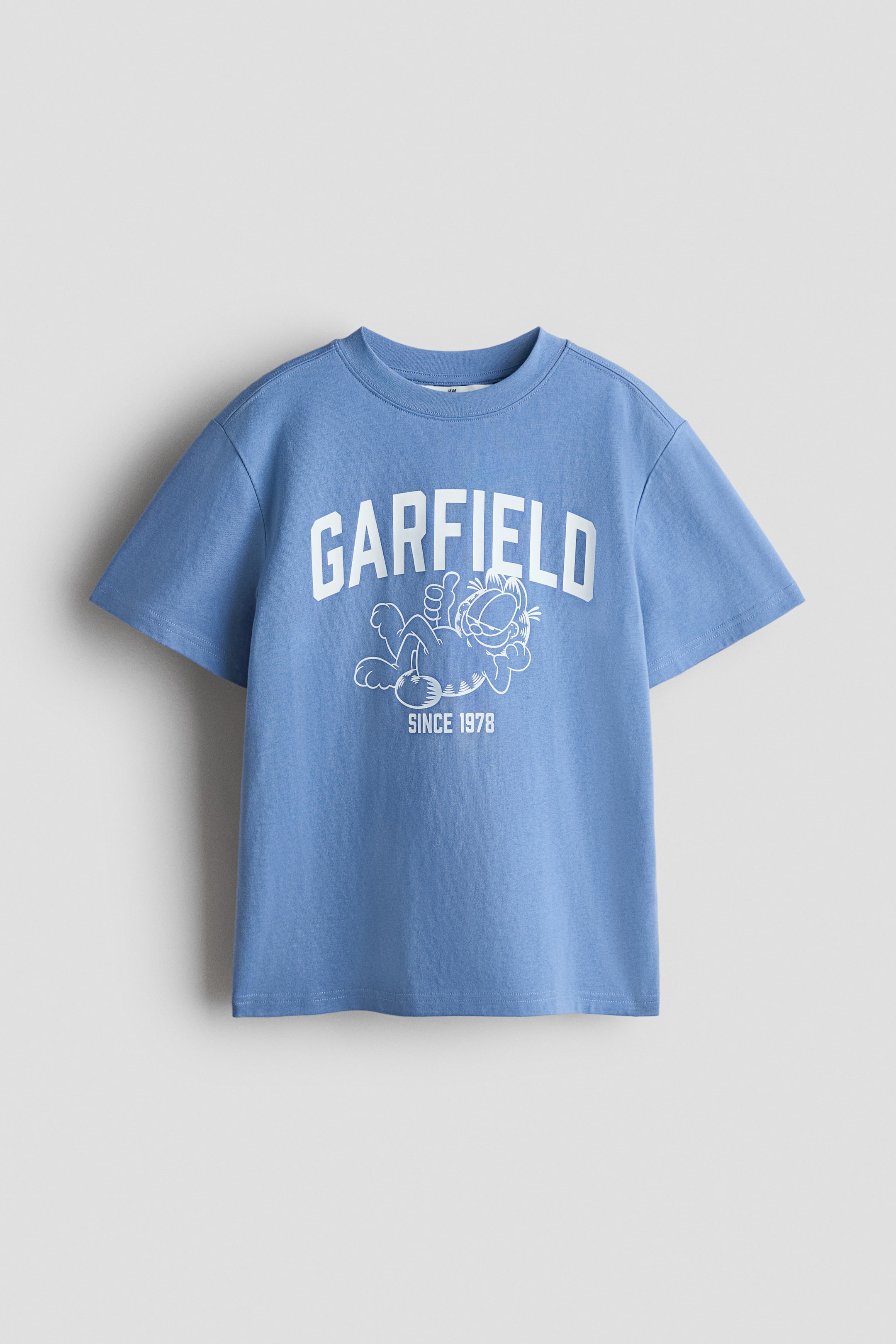 View larger image: A blue, short-sleeved t-shirt with a crew neck showcases a white graphic print on its front. The print features the word "GARFIELD" in an arched font above an outline illustration of Garfield the cat lying down, with "SINCE 1978" written below.