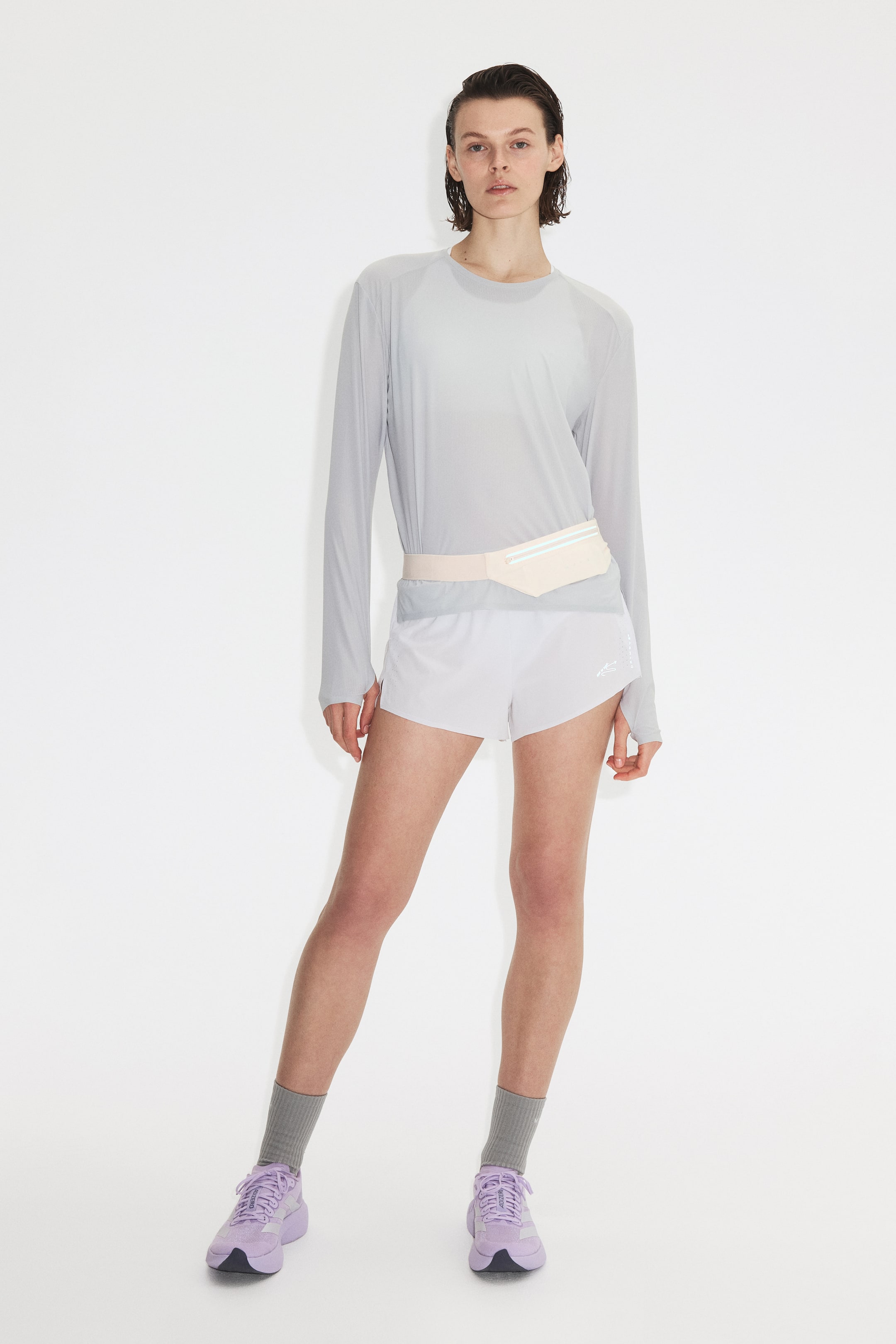 View larger image: The model is wearing a light gray, long-sleeved sports top with DryMove technology, featuring raglan sleeves and a crew neck, layered over a lighter base. A light pink fanny pack is styled at the waist over the top.