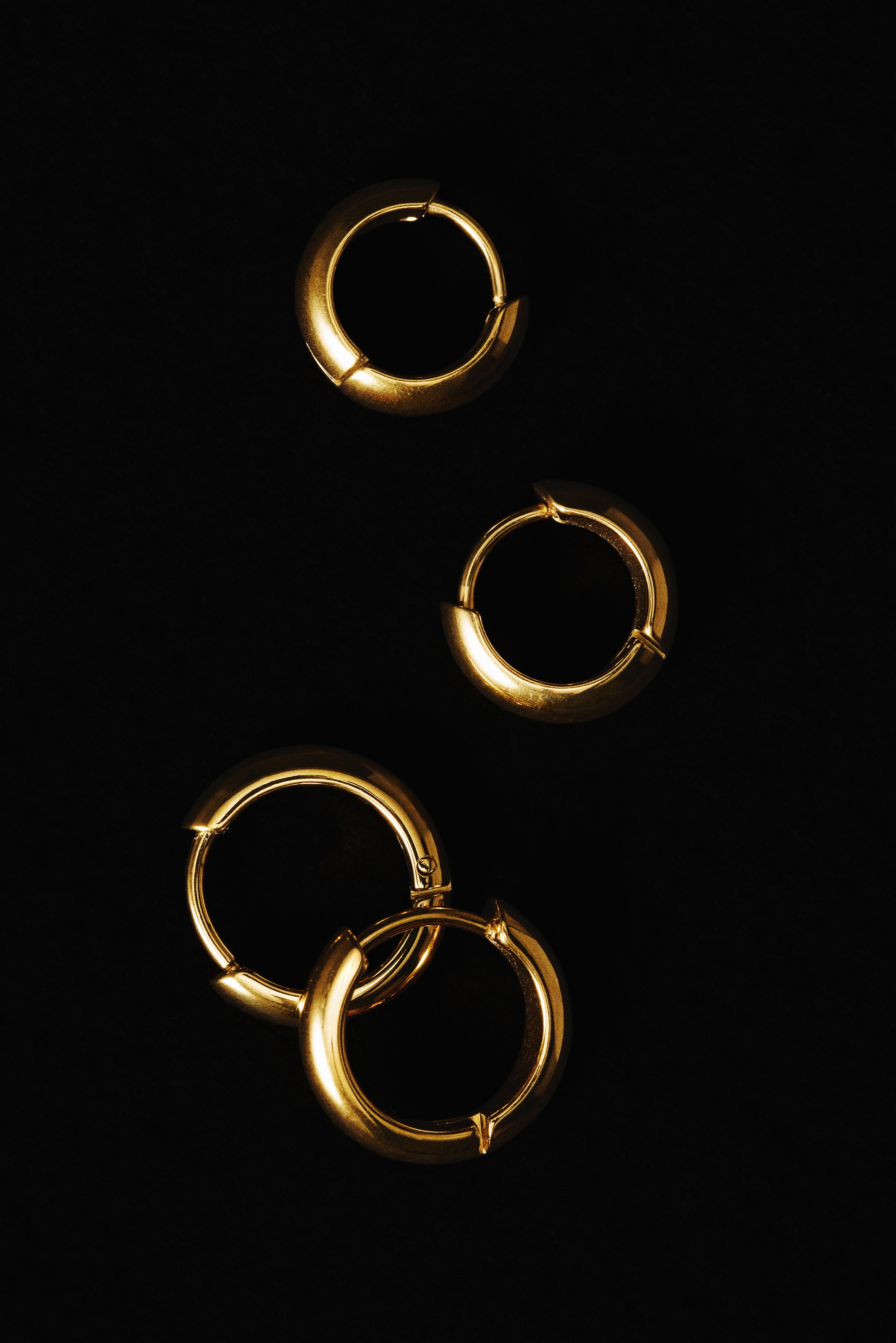 Two pairs of gold hoop earrings are displayed in a front-facing layout, arranged in a two-by-two grid. The top pair features smaller, chunky huggie hoops with a polished finish and hinged snap closures, while the bottom pair showcases a slightly larger version of the same design.