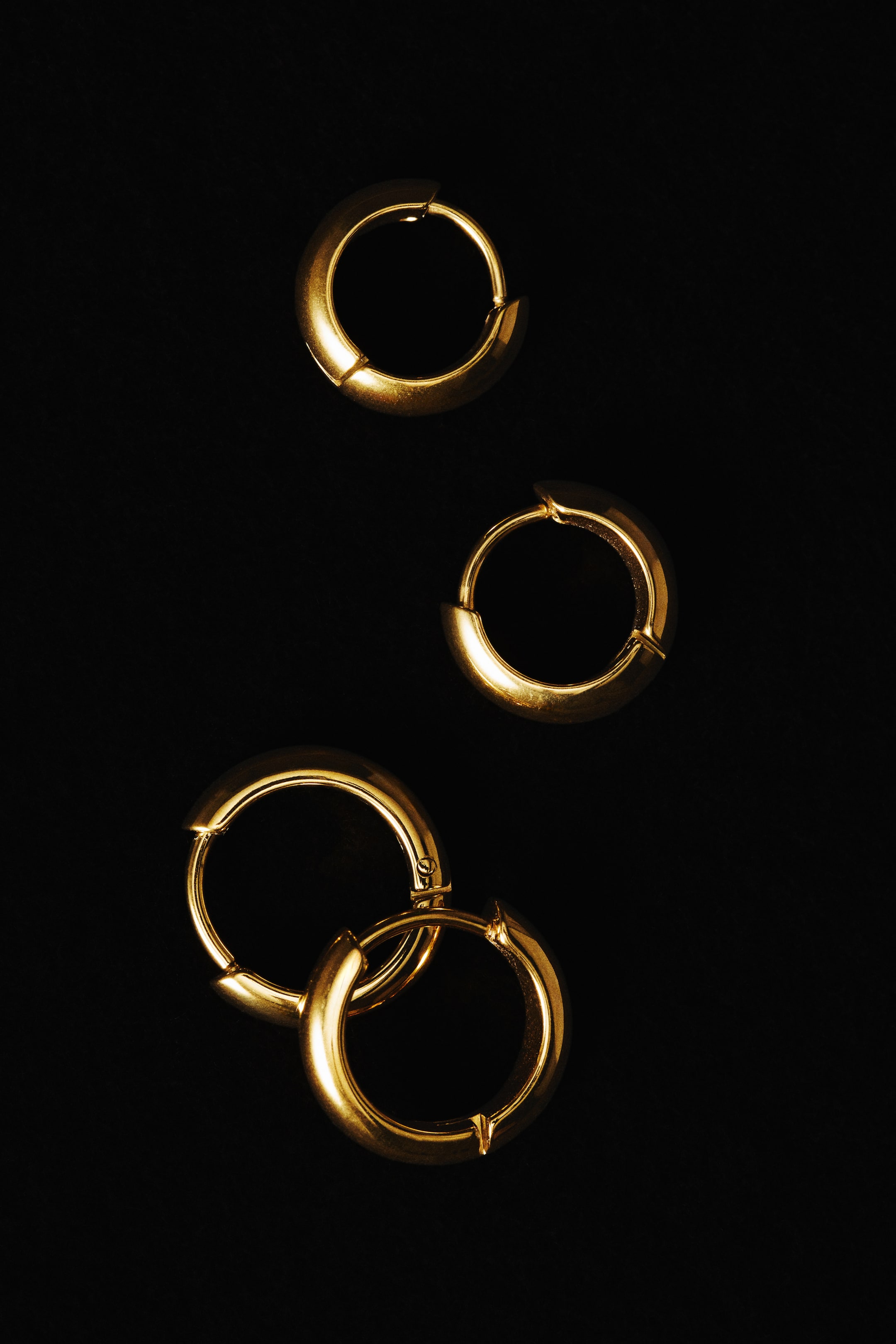 View larger image: These gold-toned stainless steel hoop earrings feature a chunky, rounded profile with a highly polished finish and integrated hinge clasps. The four hoops are shown from a front-facing view, with two individual earrings positioned above two overlapping ones.