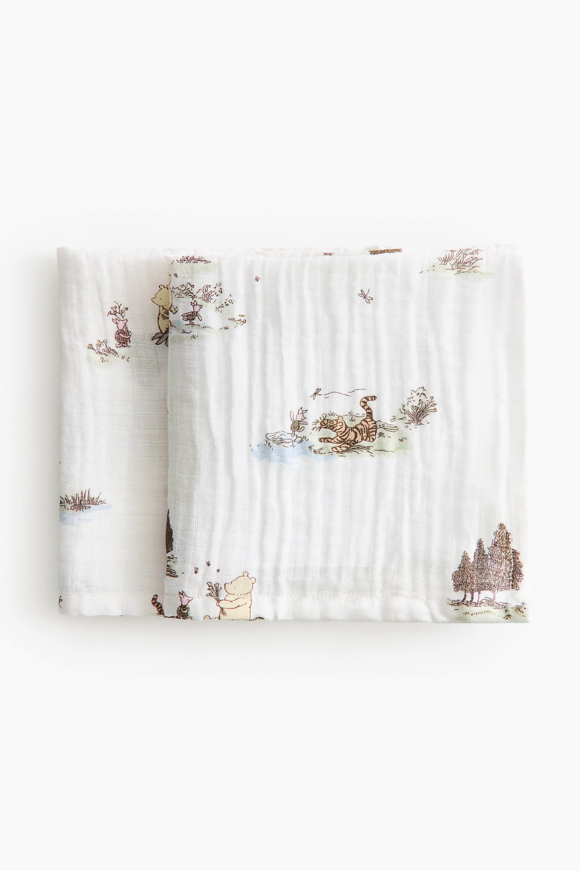 A white, crinkled muslin blanket, featuring a printed pattern of trees and cartoon characters, is draped over a light green and white gingham-checked quilted mat.
