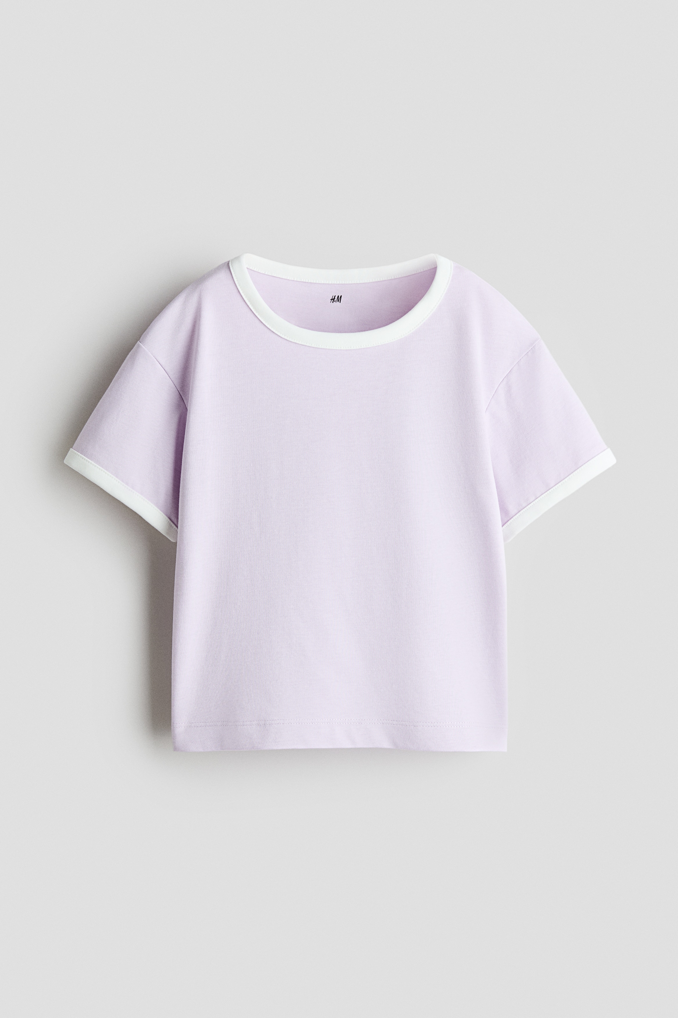 A light purple, short-sleeved ringer t-shirt with contrasting white trim on the crew neck and cuffs is worn by the model, paired with matching light purple shorts. The model is posing with crossed arms while resting their chin on their hand.