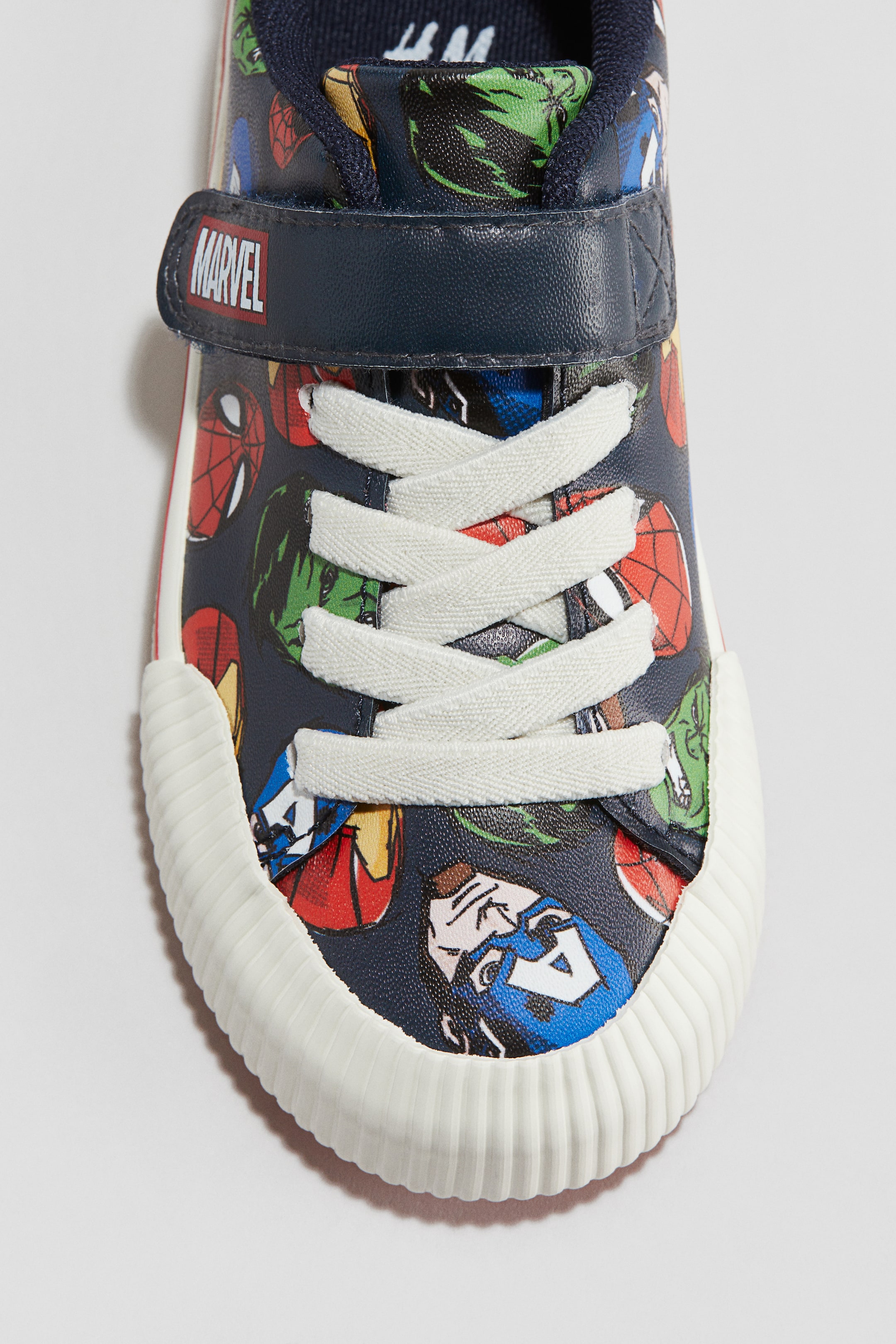 View larger image: A FRONT-facing trainer is showcasing a dark blue upper, which is being printed with various superhero character faces. White laces are crisscrossing over the vamp, below a navy strap that is displaying a red and white 'MARVEL' logo, while a cream ribbed toe cap is completing the front of the trainer, and a thin red stripe is running along its side edge.
