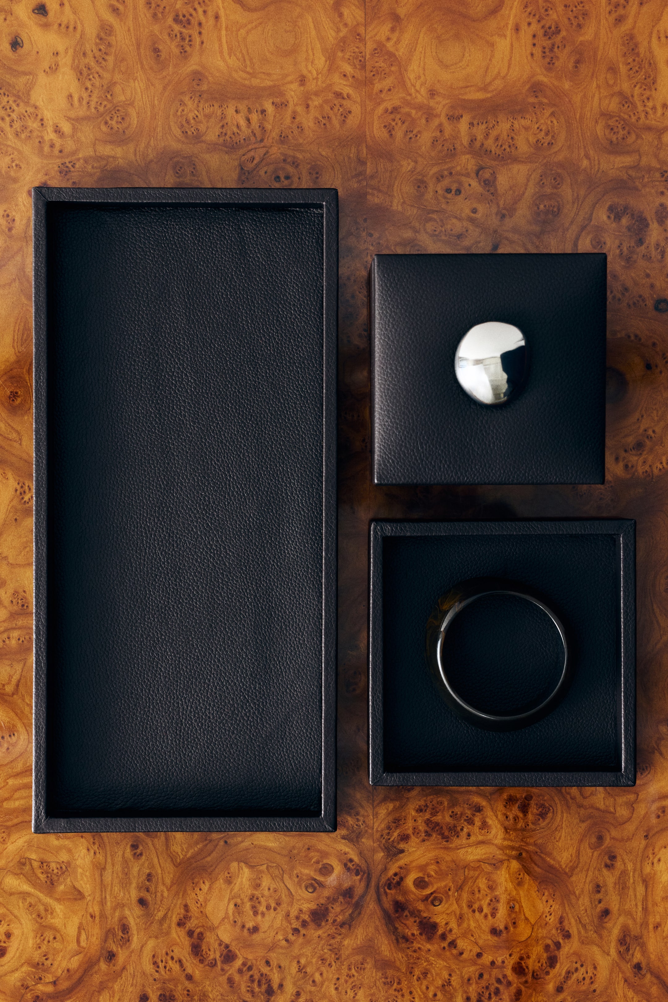 View larger image: A small, square tray covered in black textured leather is shown from a top-down perspective, featuring a low rim and holding a thick, dark circular bracelet inside. It is styled alongside a larger matching rectangular tray and a small square lidded box, showcasing its scale and design.