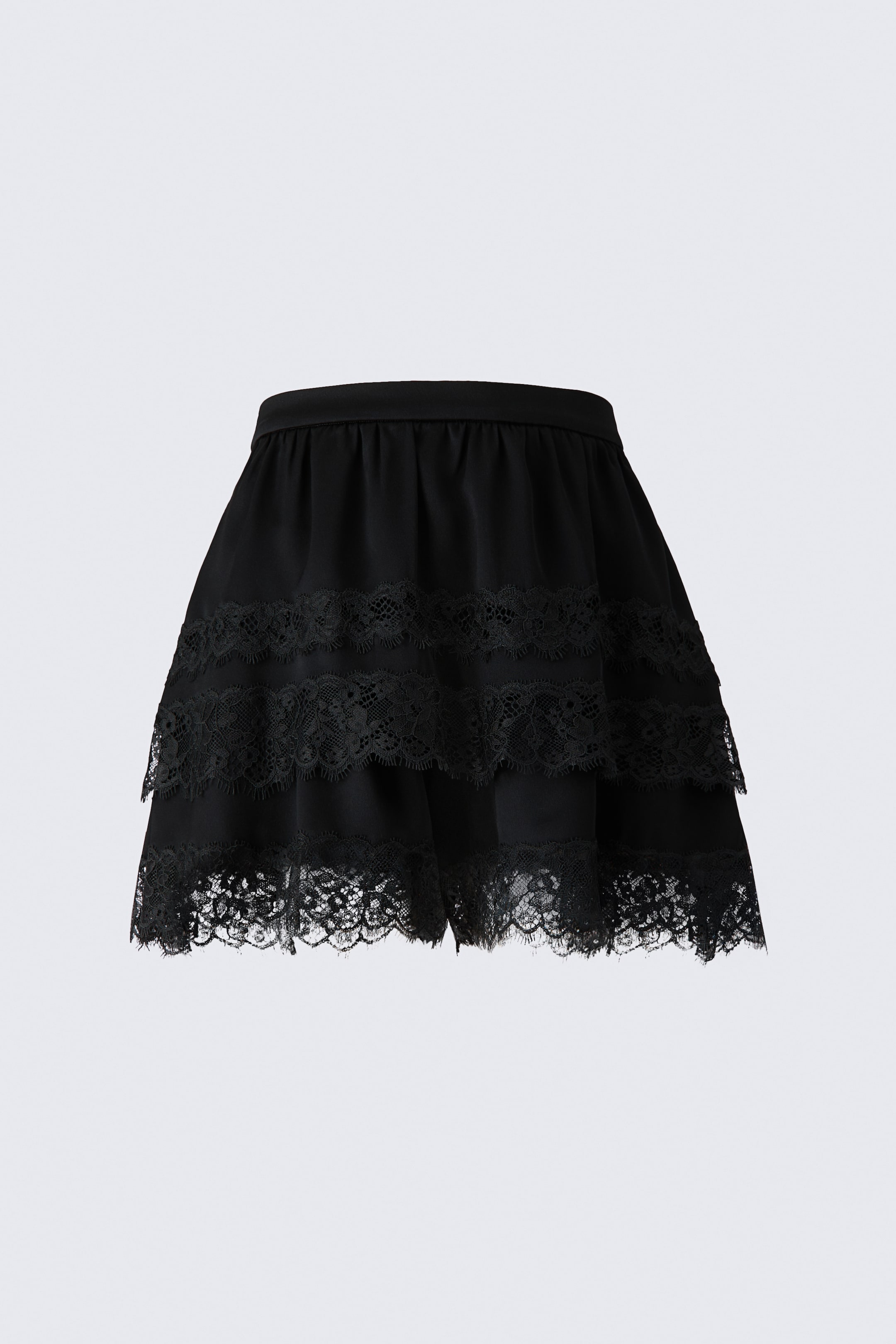 Visualizza l'immagine ingrandita: Un paio di shorts neri in raso, con design a strati effetto gonna, è mostrato da una vista frontale, con vita arricciata e silhouette svasata. Lo strato superiore presenta due bande orizzontali di rifinitura in pizzo smerlato con bordo frangiato, mentre l’orlo dello strato inferiore è rifinito con un’ampia bordura in pizzo coordinata.