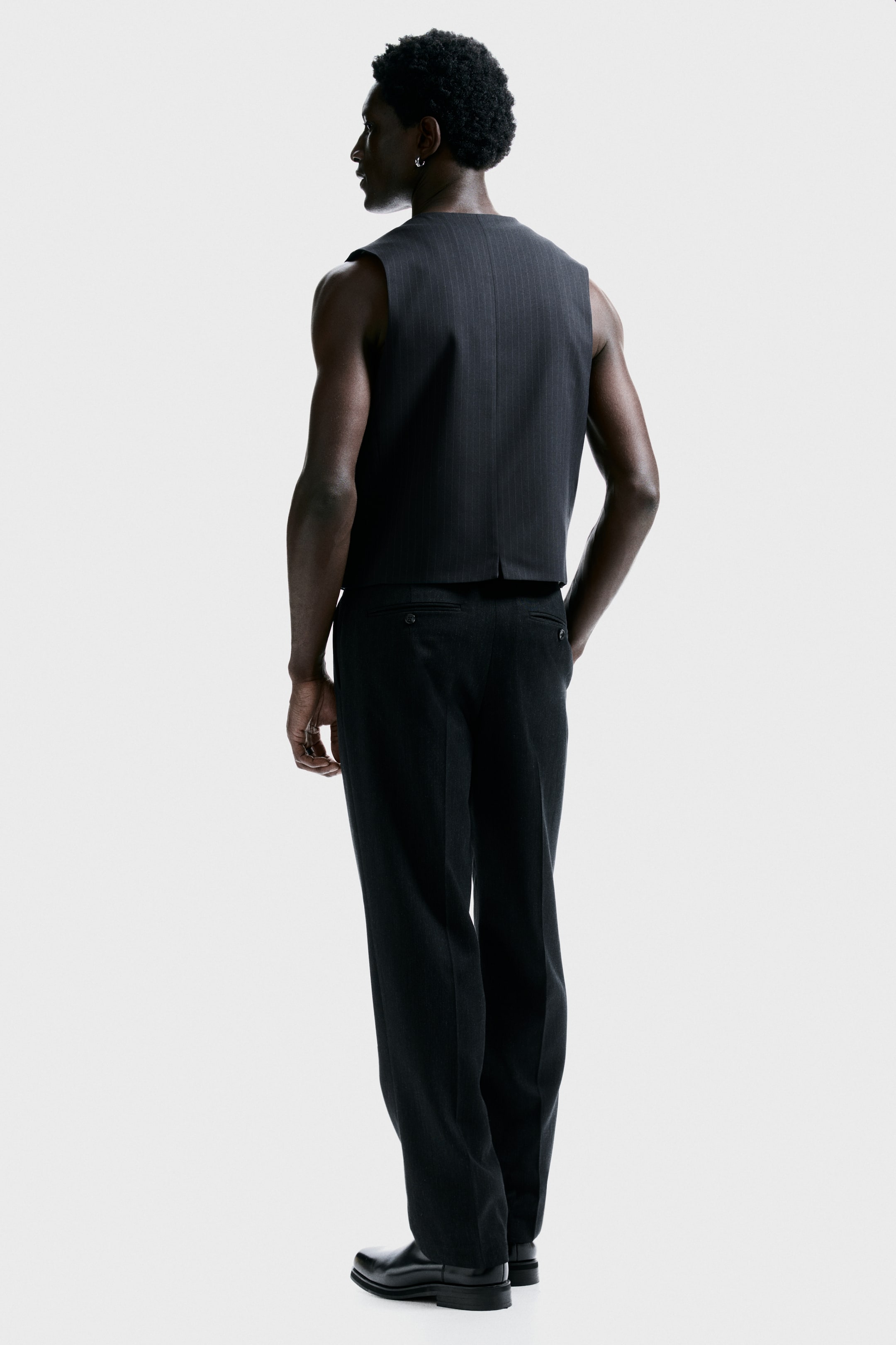 View larger image: A dark pinstriped vest is being displayed from a back-right three-quarter angle, featuring a sleeveless design, a cropped hem, and a small back vent. Matching dark trousers are being worn, presenting a relaxed fit and two buttoned back pockets.